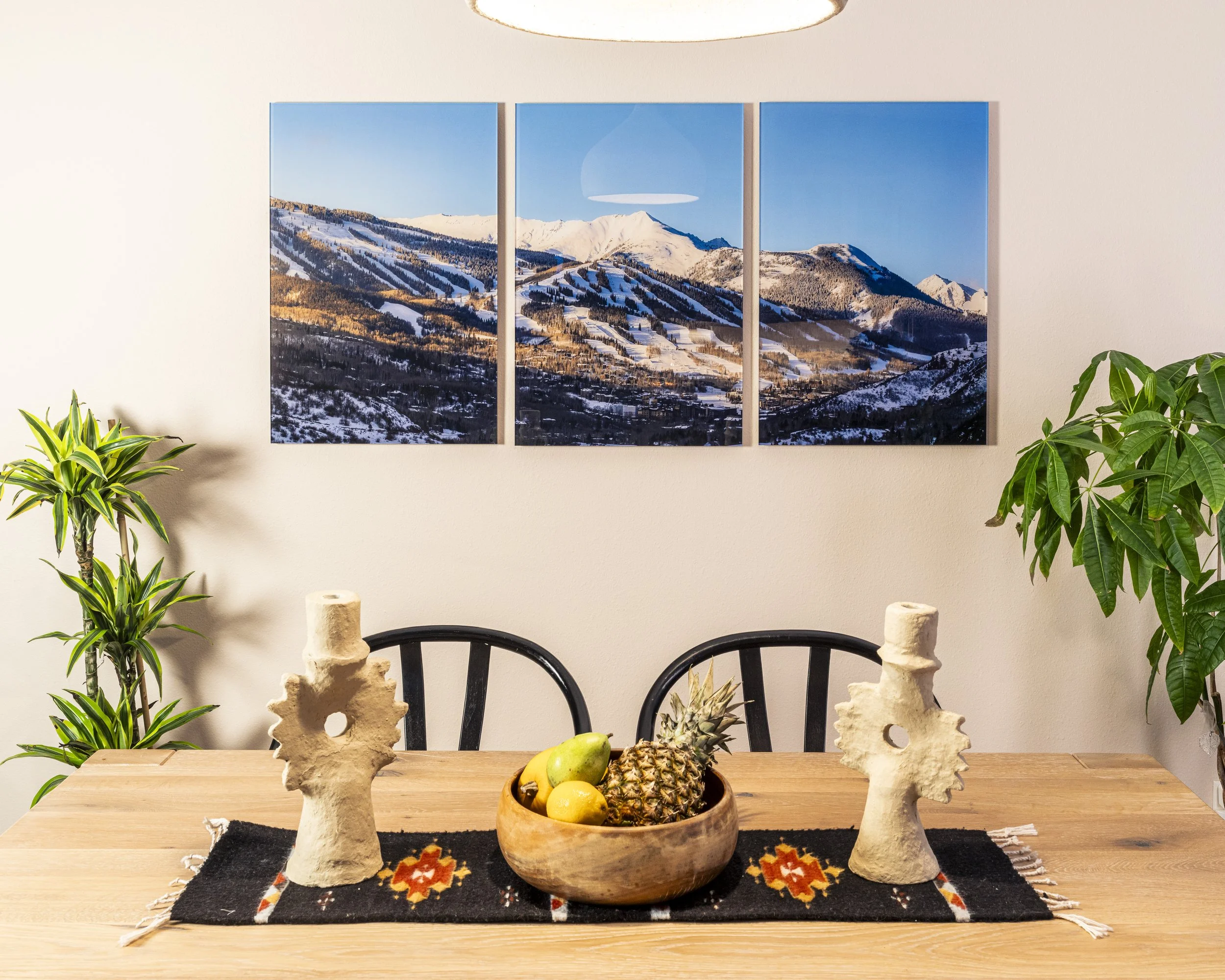 A dining table with a centerpiece of tropical fruits including pineapple, bananas, and apples, flanked by two sculptural coral-like objects, with green plants on both sides and a triptych of mountain landscape art on the wall in the background.
