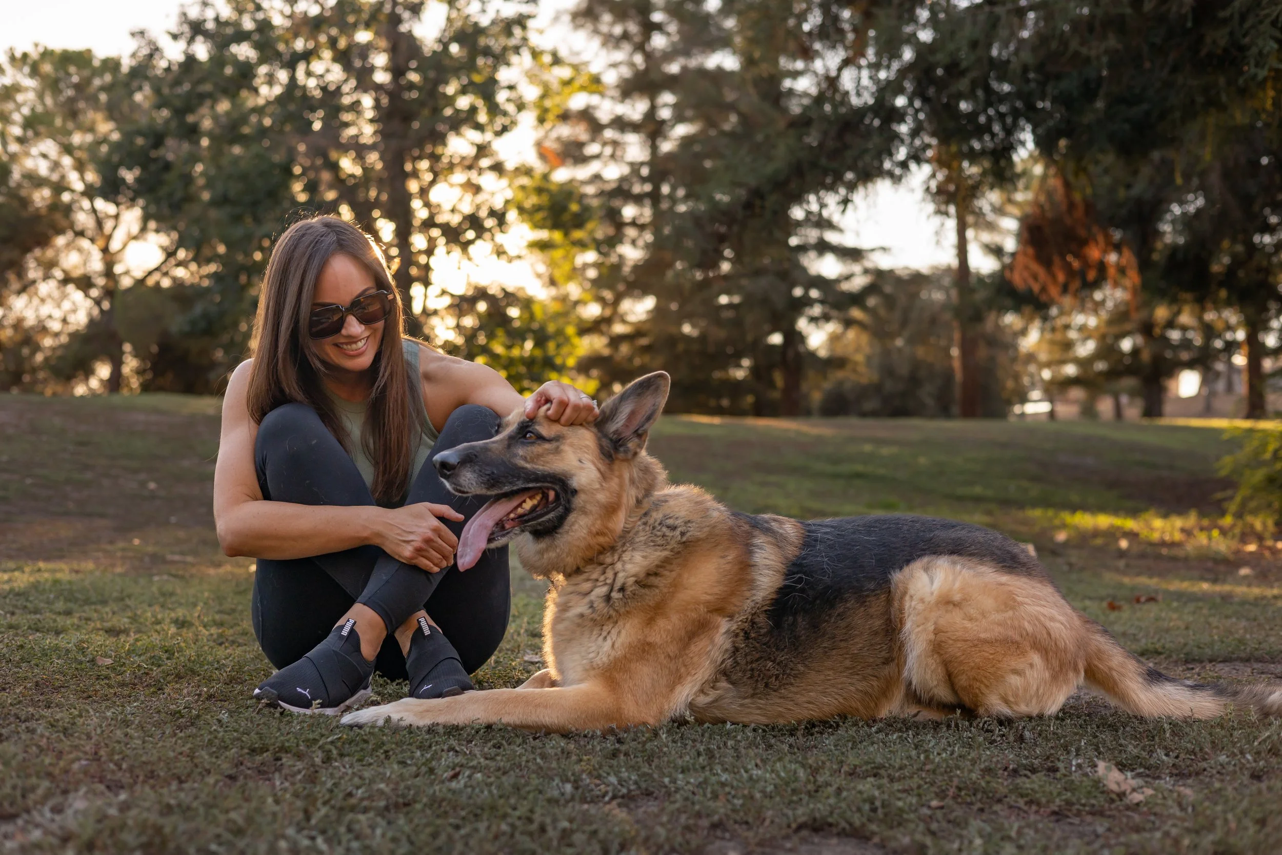 A smiling woman wearing sunglasses sitting on the ground petting a large German Shepherd dog lying down on the grass in a park during sunset.
