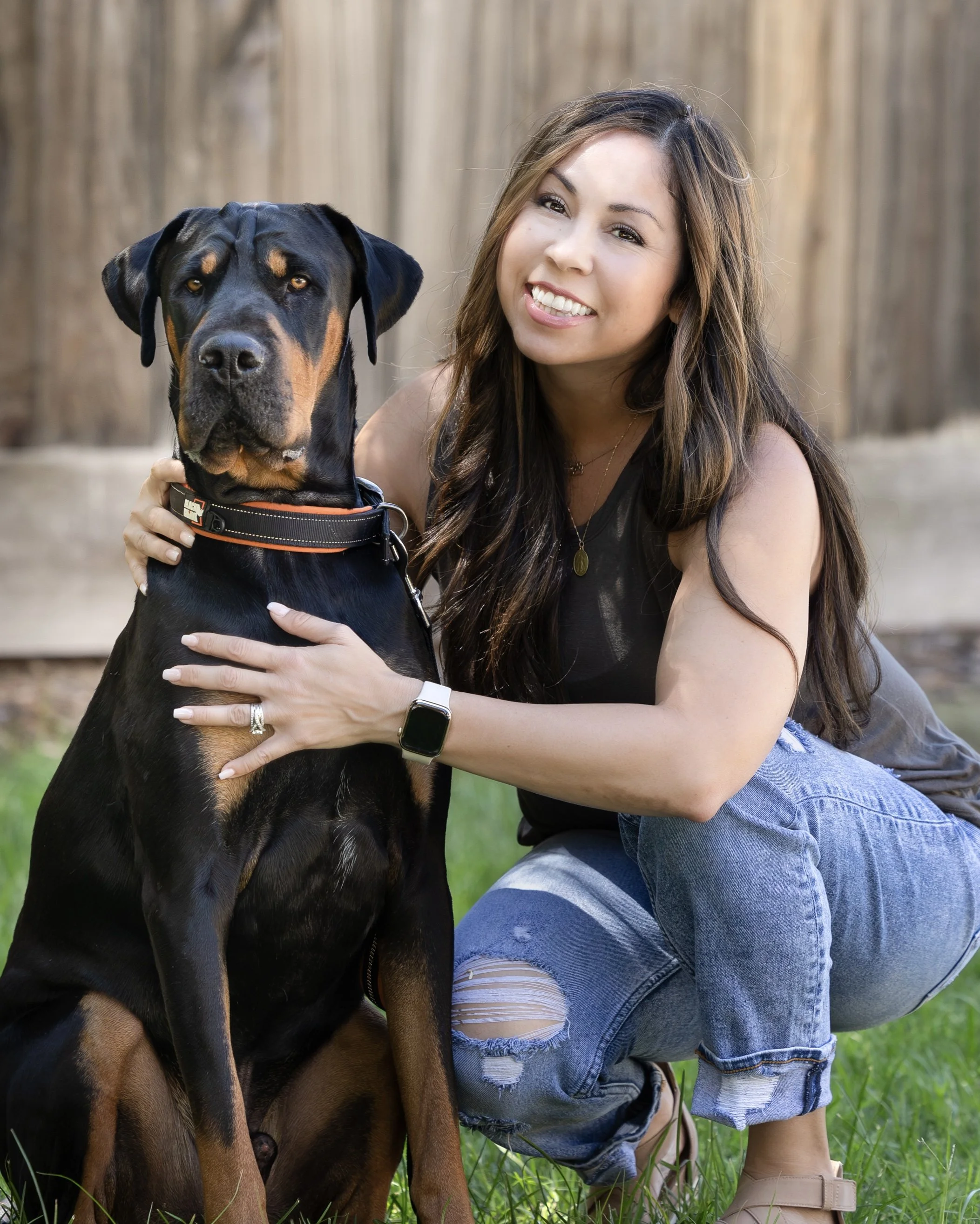 A woman with long brown hair, wearing a sleeveless black top and ripped jeans, kneeling outdoors on green grass, smiling and holding a large black and tan Doberman dog with a collar.