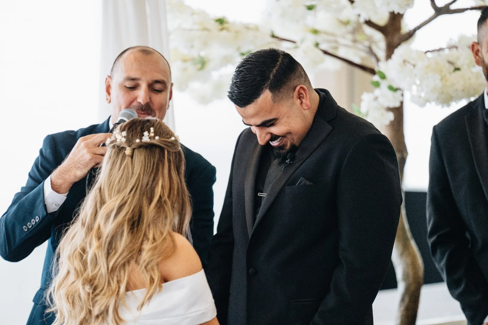 A wedding ceremony with a bride and groom exchanging vows, Chris Bragg holding a microphone, and floral décor in the background.