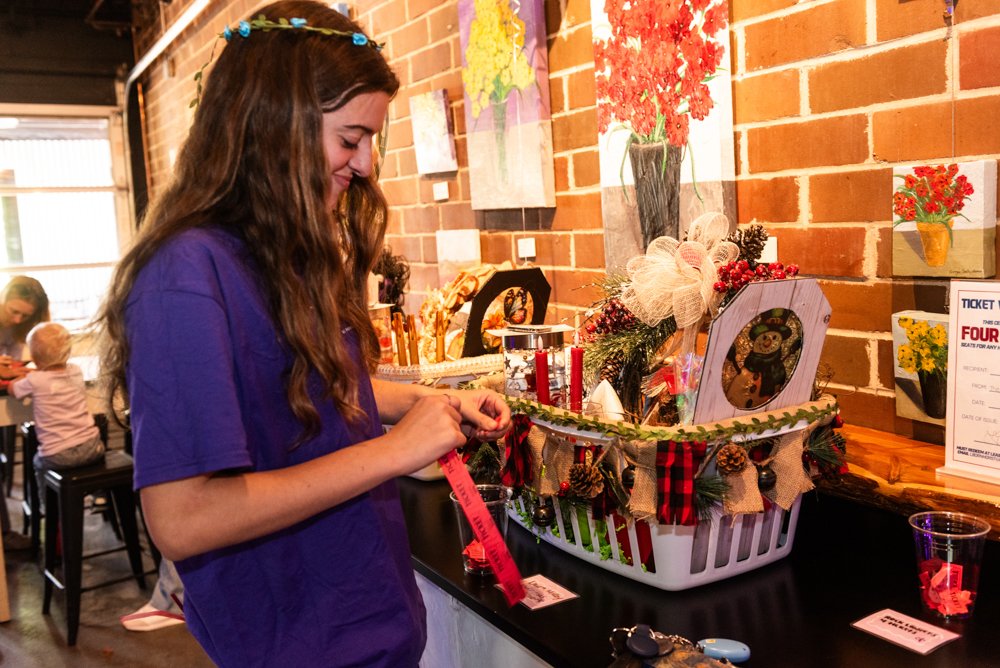 Young woman with long brown hair, wearing a purple shirt and a blue floral headband, arranging items in a holiday-themed gift basket on a table inside a brick-walled room with artwork and other people in the background.
