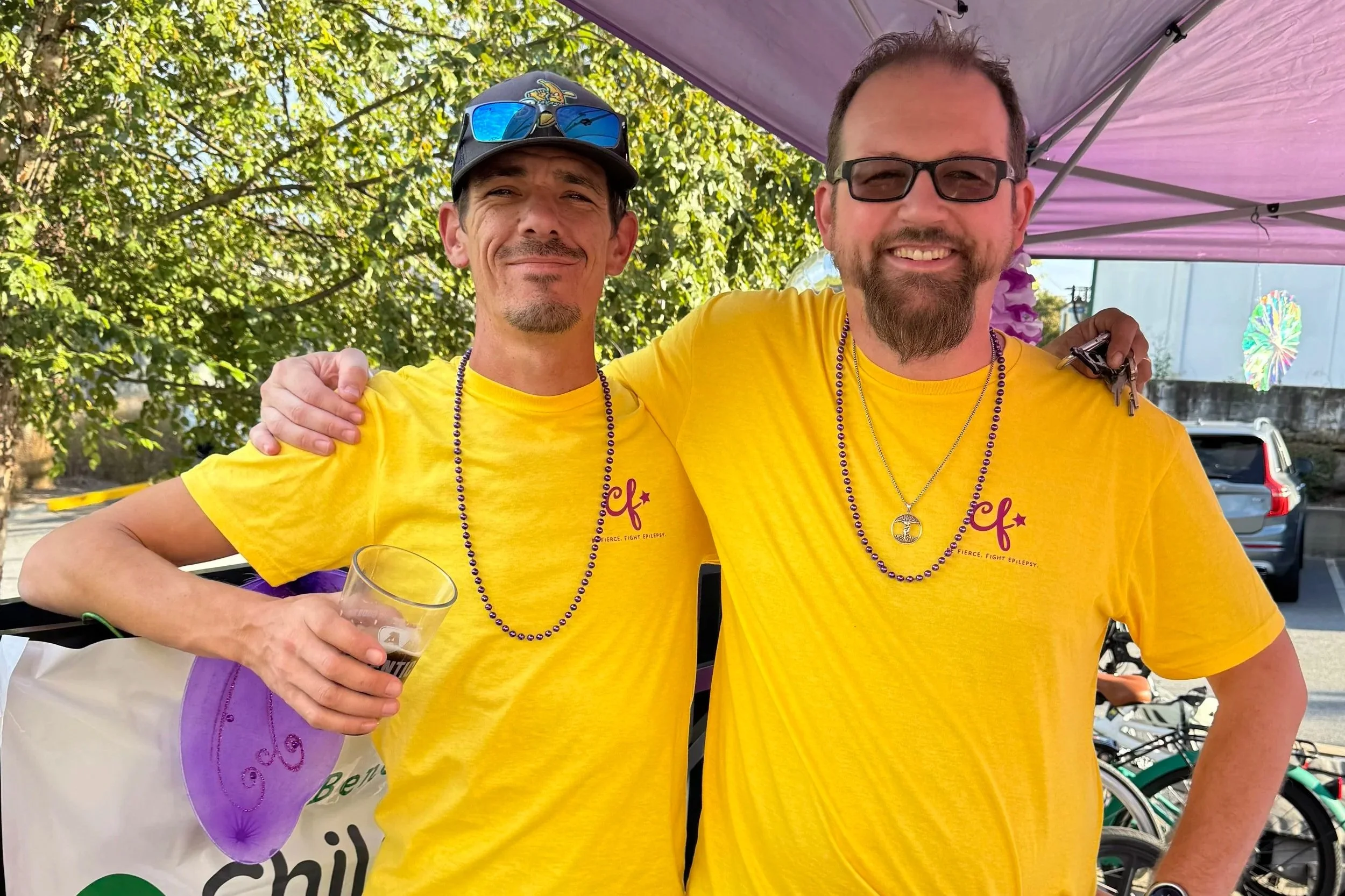 Two men wearing yellow T-shirts standing outdoors under a pink canopy, smiling, with trees and parked cars in the background. One man has sunglasses on his cap and the other wears glasses, both wearing beaded necklaces.