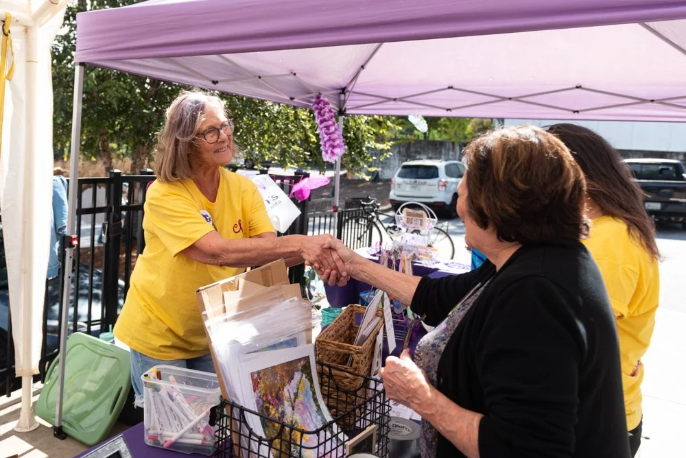 A woman in a yellow shirt shaking hands with an elderly woman at an outdoor booth under a purple canopy, with various items on the table and parked cars in the background.