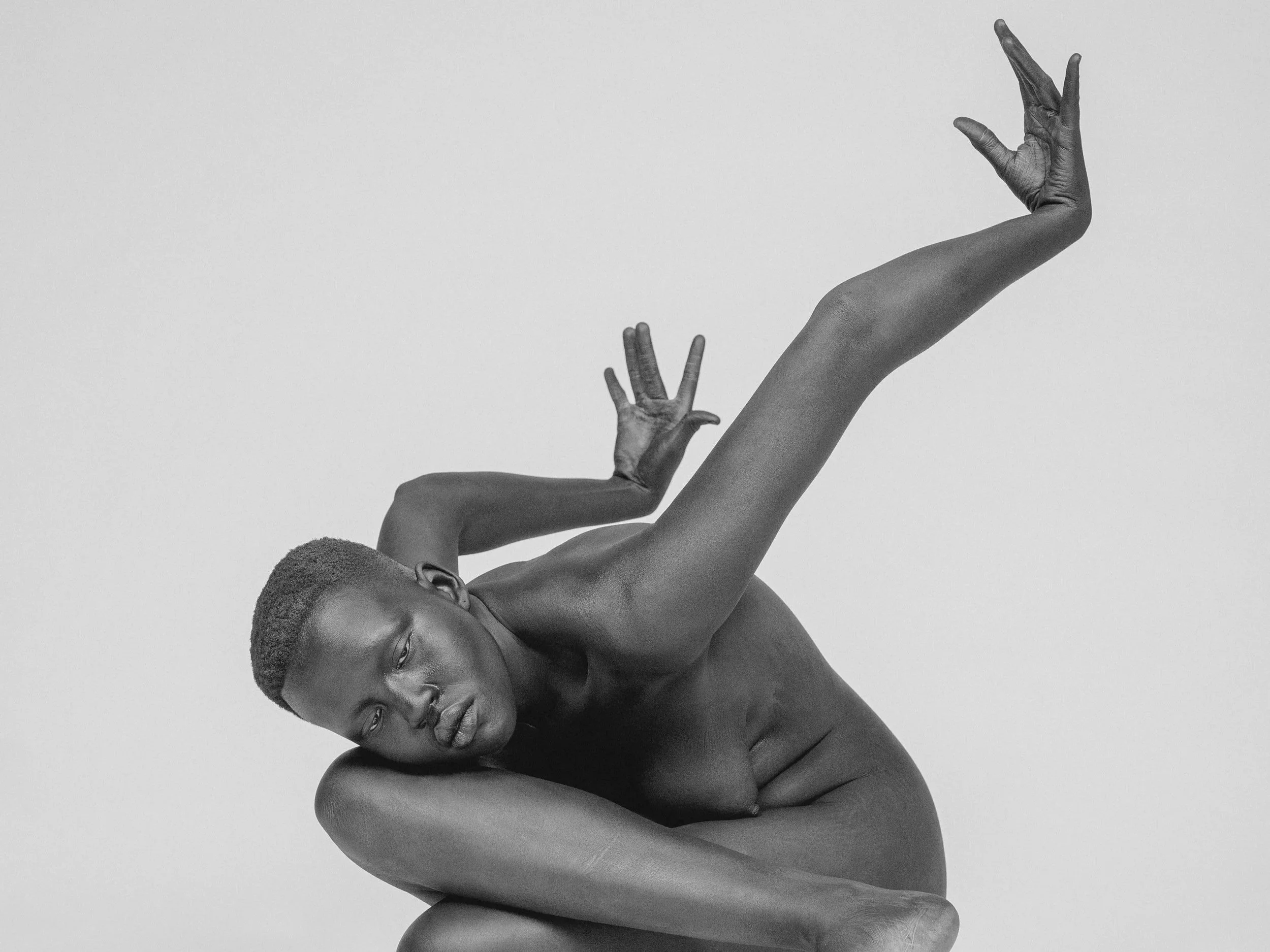 A black and white photo of a topless woman with short hair, with one arm bent behind her head and the other arm raised with fingers stretched out.