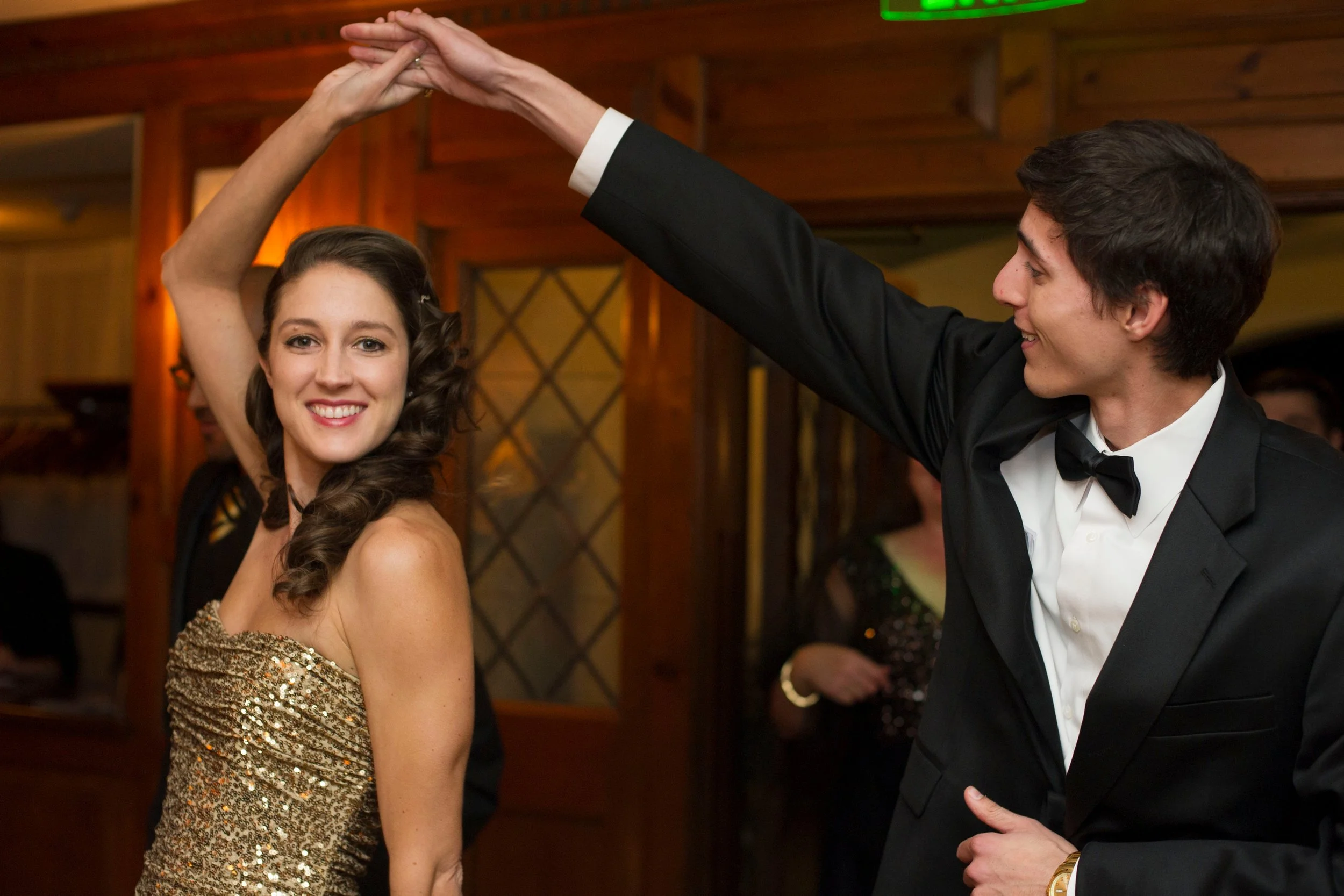 A young woman and a young man in formal attire dancing together at a party or wedding reception. The woman is smiling and wearing a sparkling gold dress, and the man is wearing a black tuxedo with a bow tie. They are holding hands in a dance pose, with a wooden interior setting in the background.