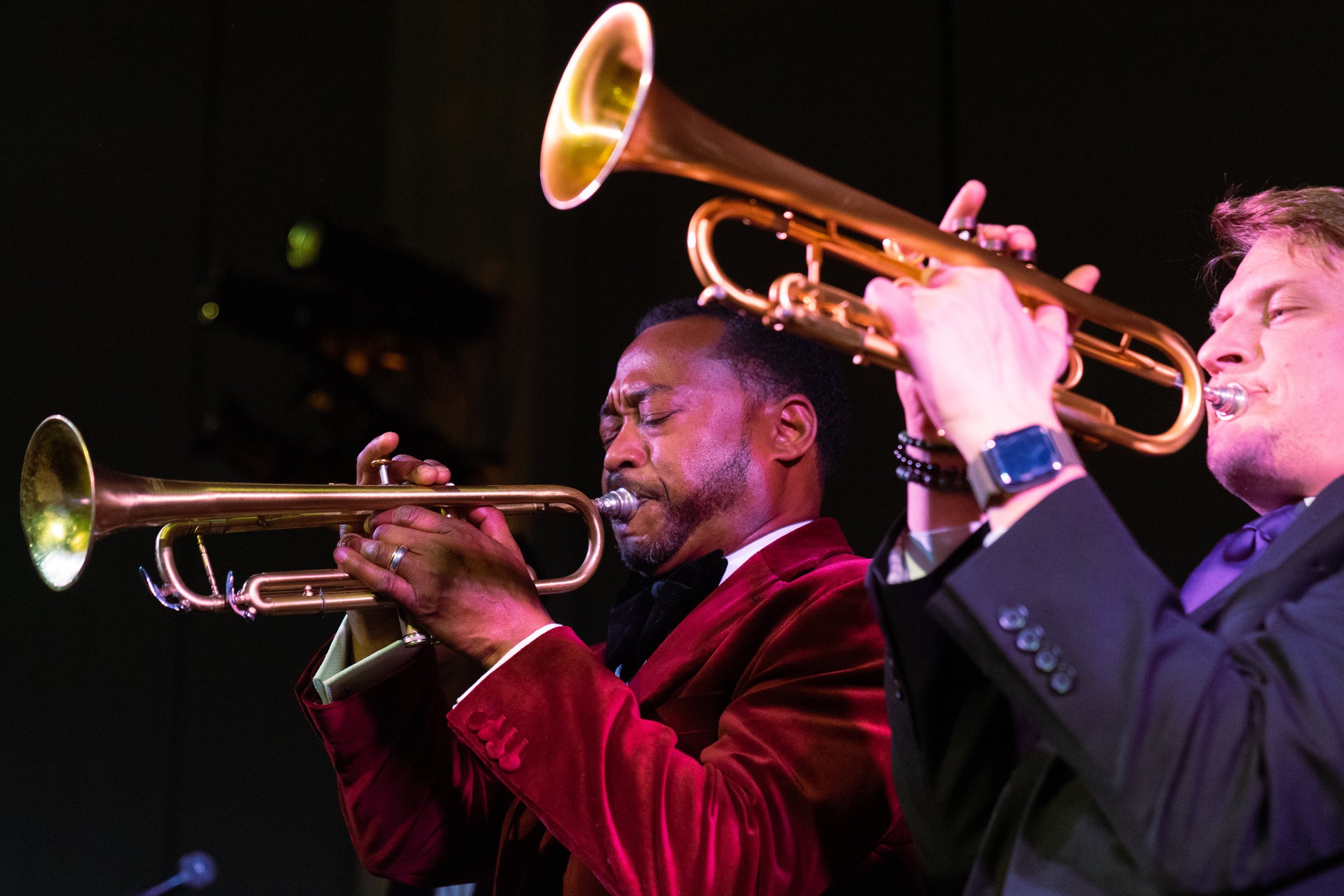 Two men in tuxedos playing trumpets on stage with a dark background.