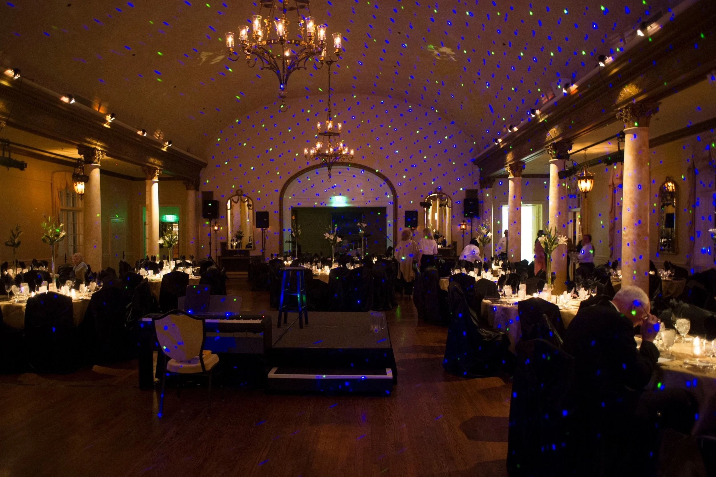 Elegant banquet hall decorated for a formal event with round tables, tall centerpieces, chandeliers, and wall mirrors, illuminated with dim lighting and colorful laser lights projected on the ceiling and walls.