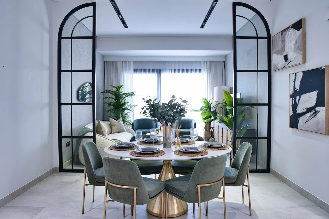 A modern dining room with a round table set for six, decorated with black plates, gold chargers, and glassware. The table has a gold pedestal base and a large central floral arrangement. Behind the dining area, there's a bright living room with a beige sofa, green armchair, and large potted plants, separated by black metal-framed glass partitions, with light-colored curtains and abstract artwork on the walls.