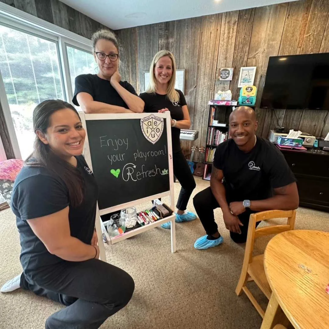 Four people, three women and one man, smiling and posing in a playroom with a chalkboard that says 'Enjoy your playroom Refresh.' The room has wooden walls, a window, a small wooden table, and shelves with books and toys.