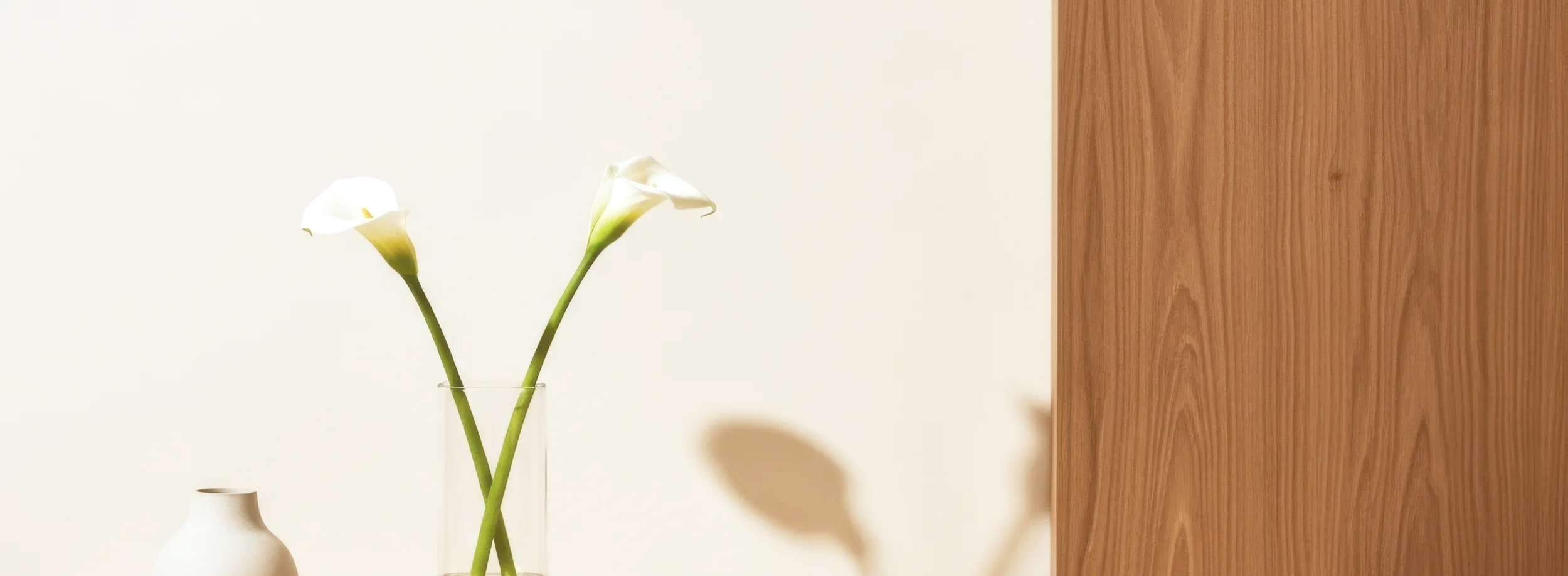 A minimalist interior with two white calla lily flowers in a clear glass vase and a small white ceramic vase on a surface, against a plain wall and a wooden panel.