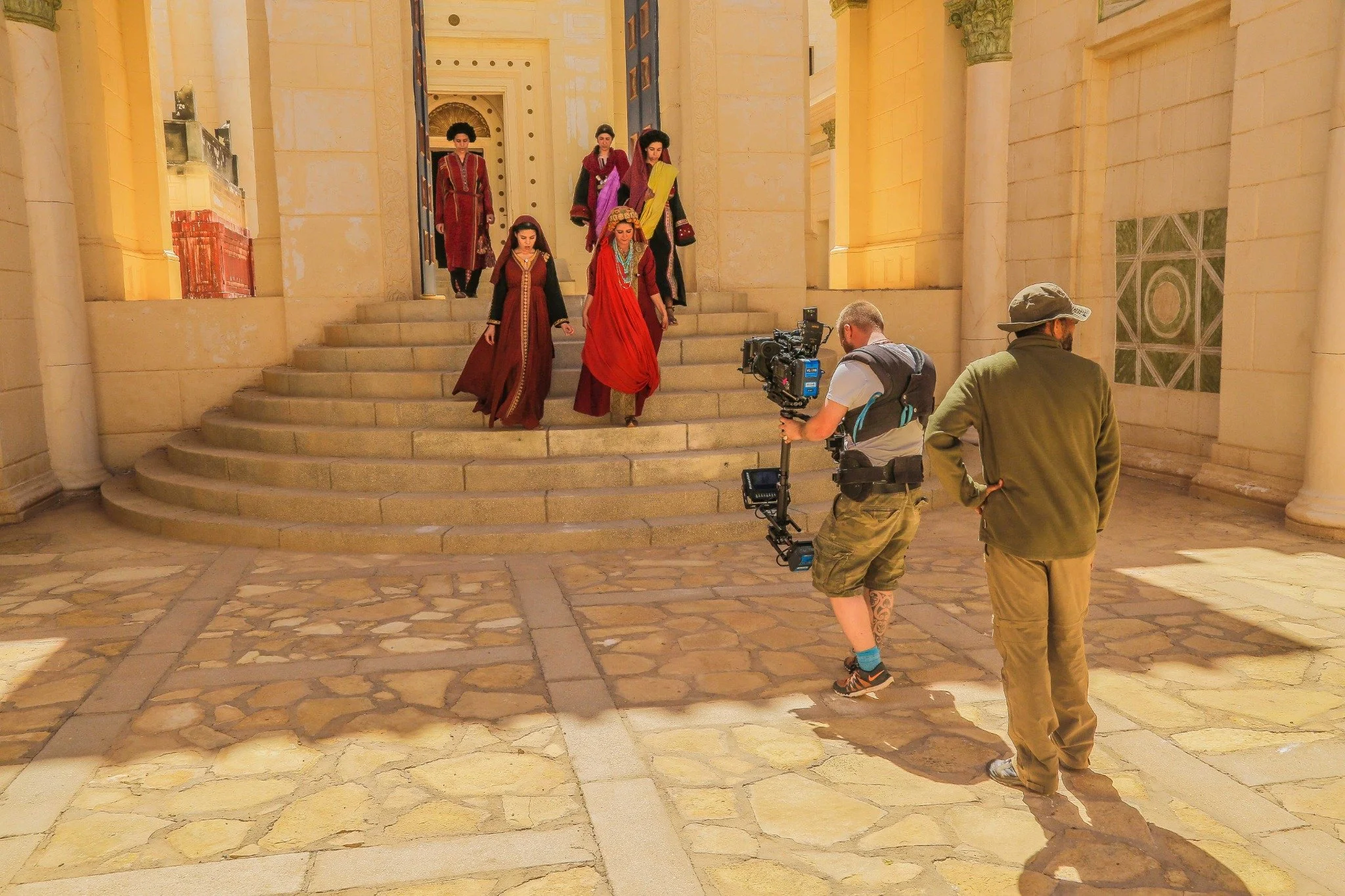 Costumed cast descending palace steps with a camera operator on a film set in Morocco.