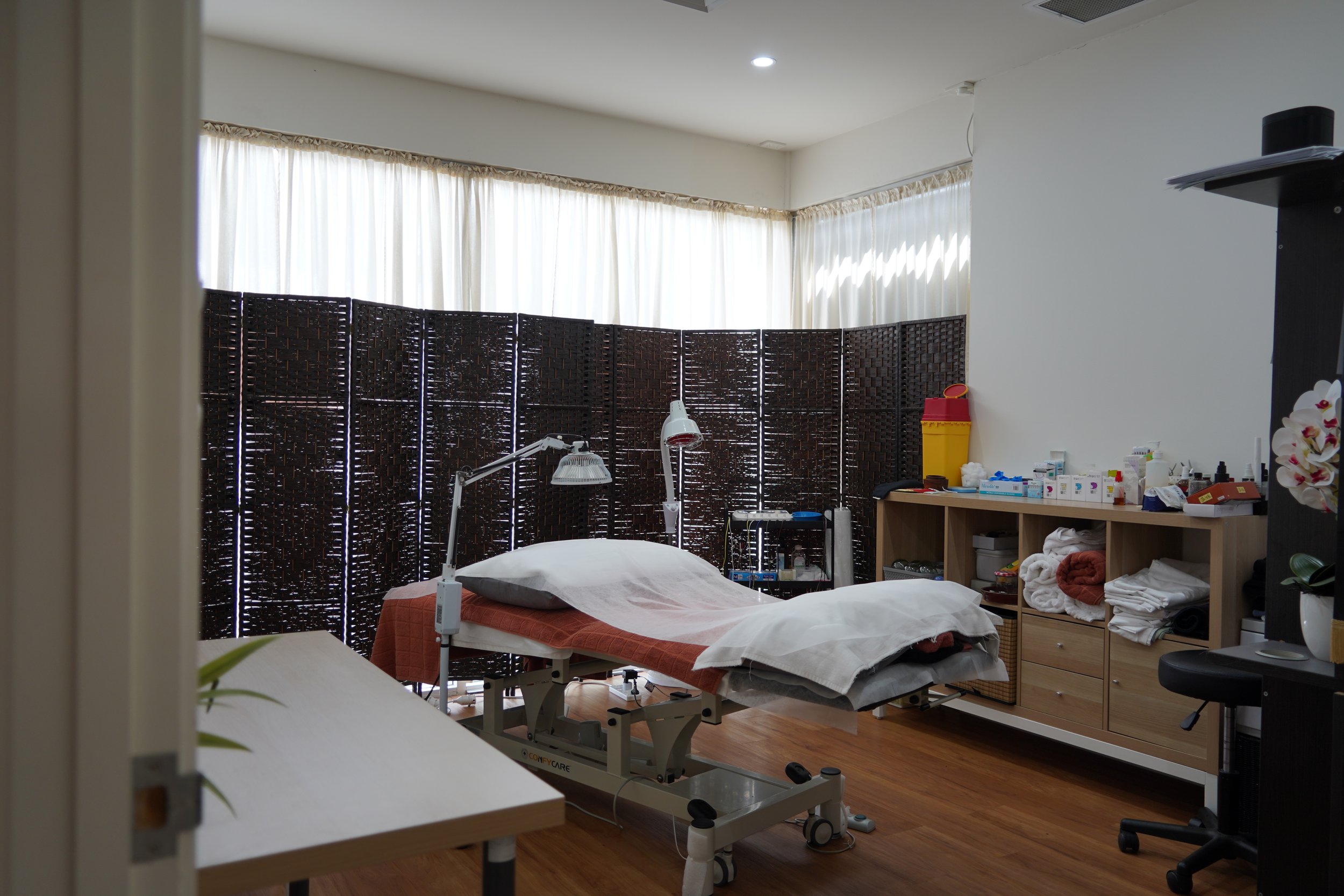 A medical examination room with a treatment bed, folded towels, medical supplies on shelves, and a privacy screen in the background.