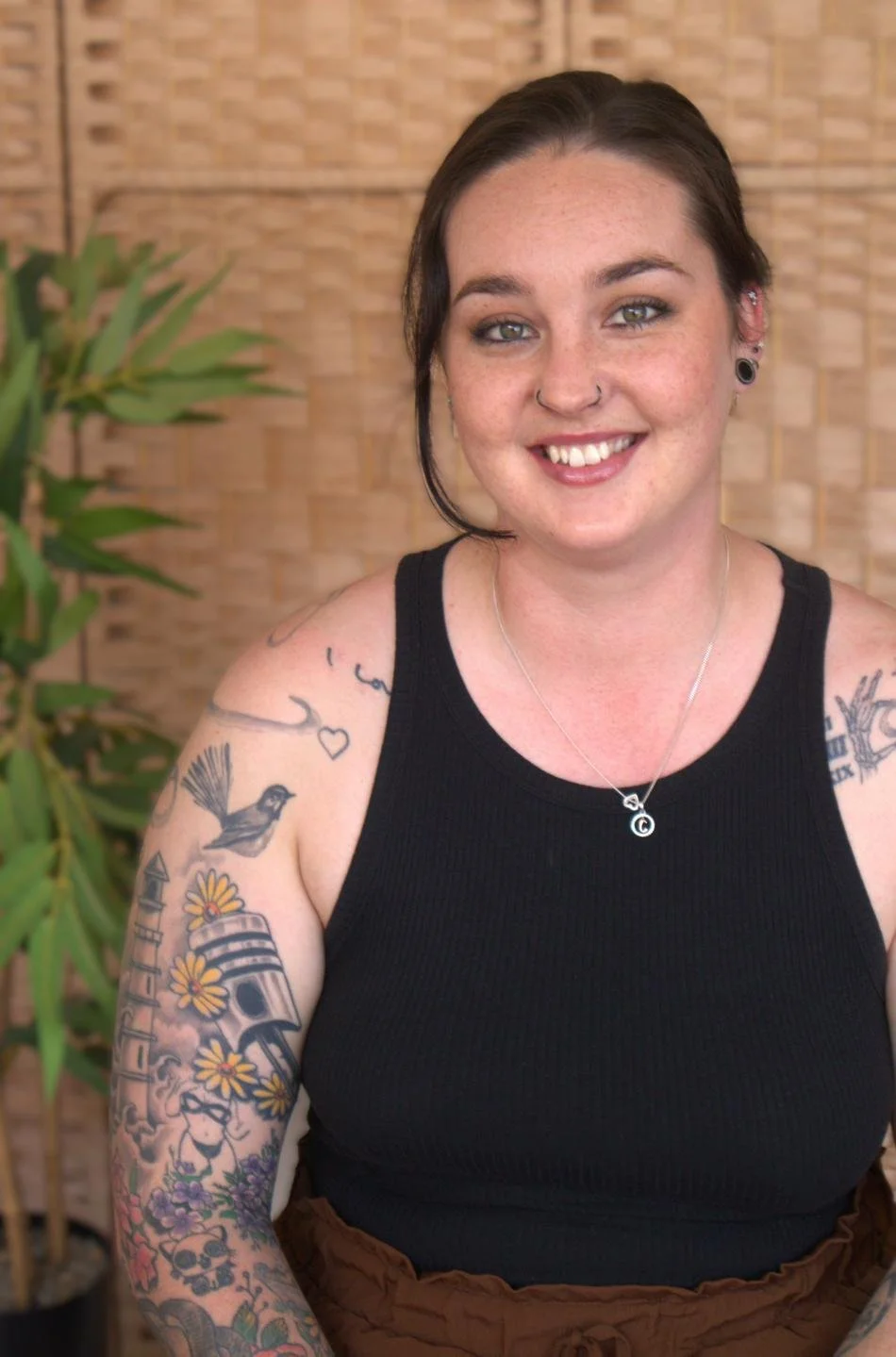 Young woman with dark hair, red lipstick, and a nose piercing, smiling outdoors with a blurred natural background.