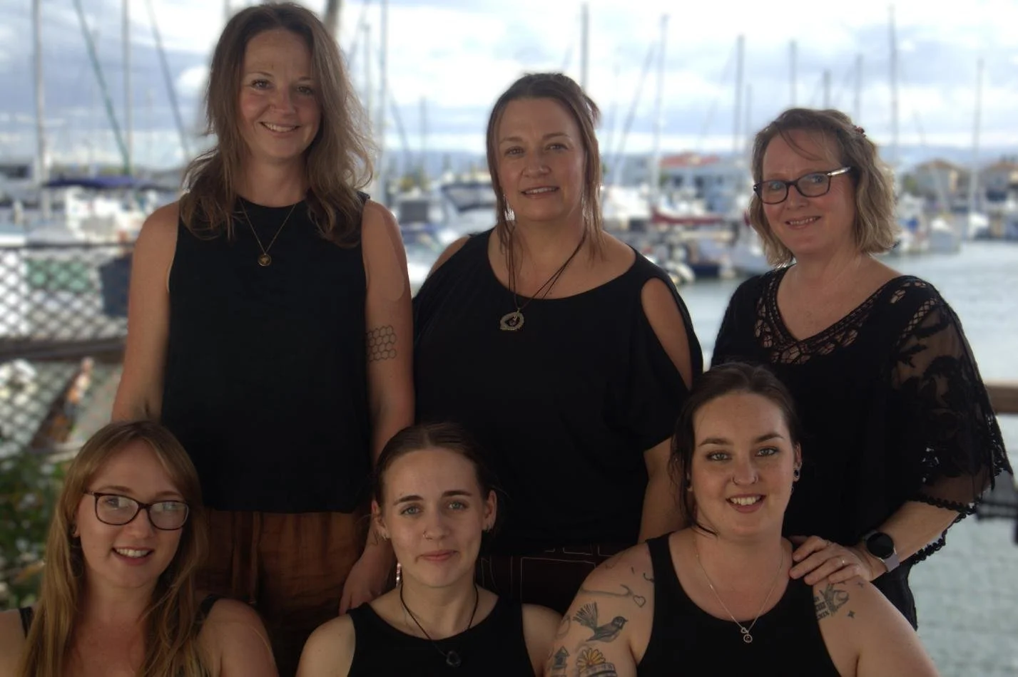 Six women posing together at a marina with boats in the background, all wearing black tops.
