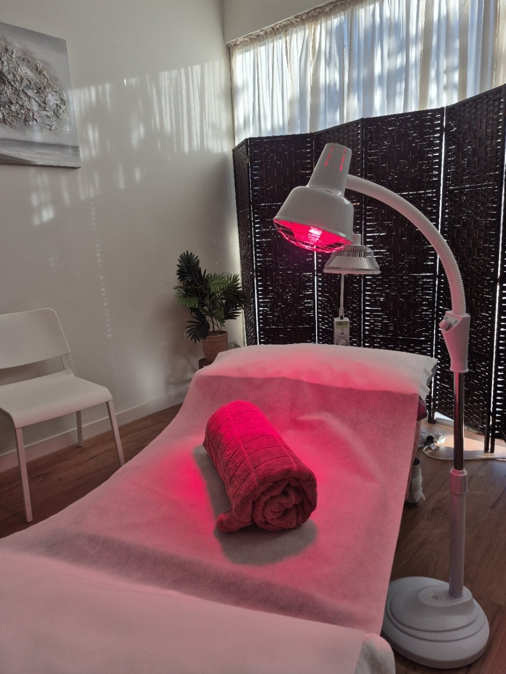 A massage or facial treatment room with a massage table covered in a white sheet, a folded red towel on it, a tall floor lamp with a red light, a black privacy screen, a small potted plant, and a white chair in the corner.