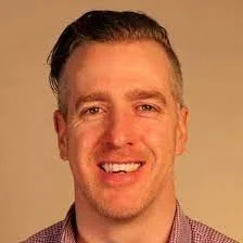 Portrait of a smiling man with short, dark hair, wearing a collared shirt, against a beige background.
