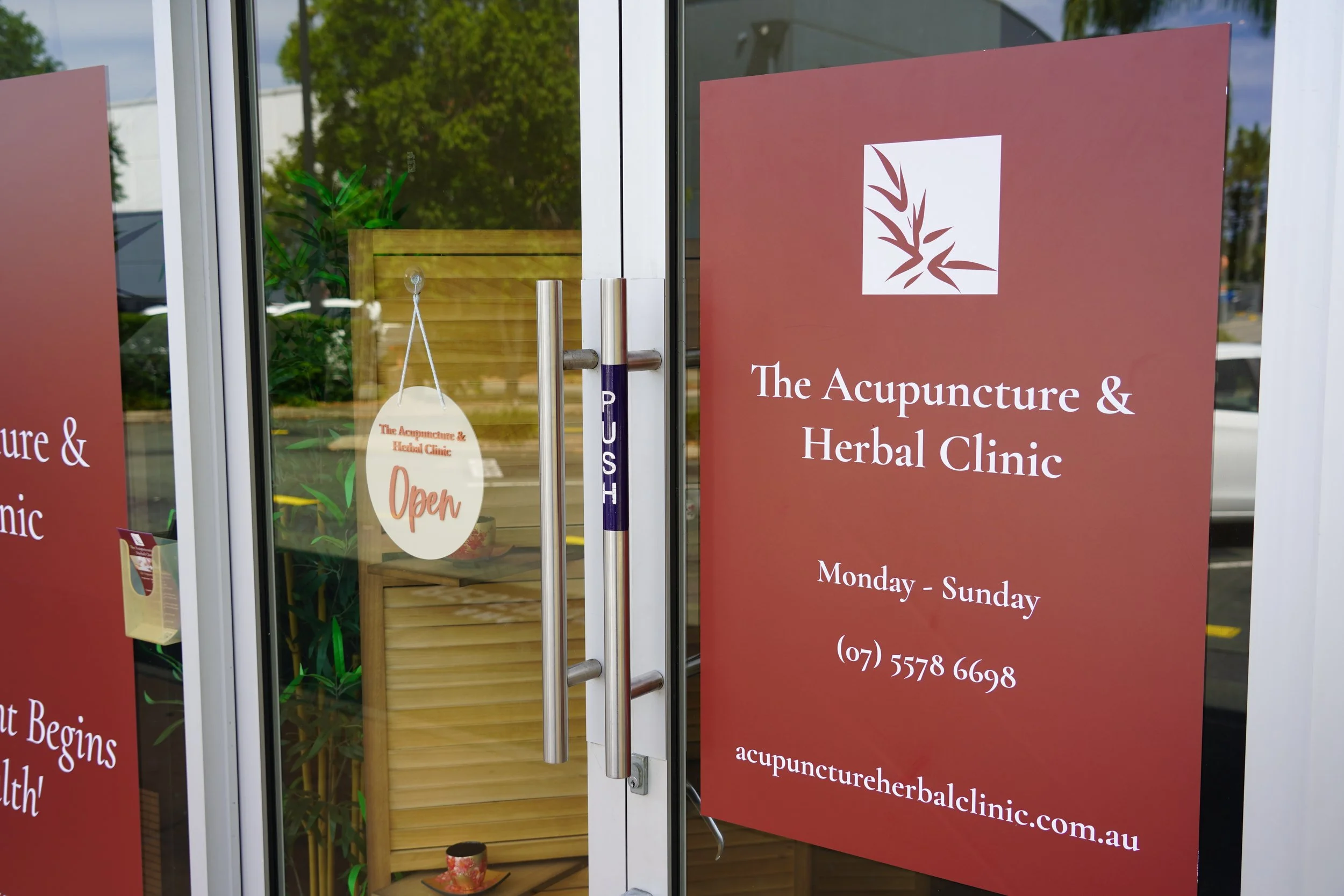 The entrance of The Acupuncture & Herbal Clinic with a large red sign displaying the clinic's name, hours, phone number, and website, and an 'Open' sign hanging on the glass door.