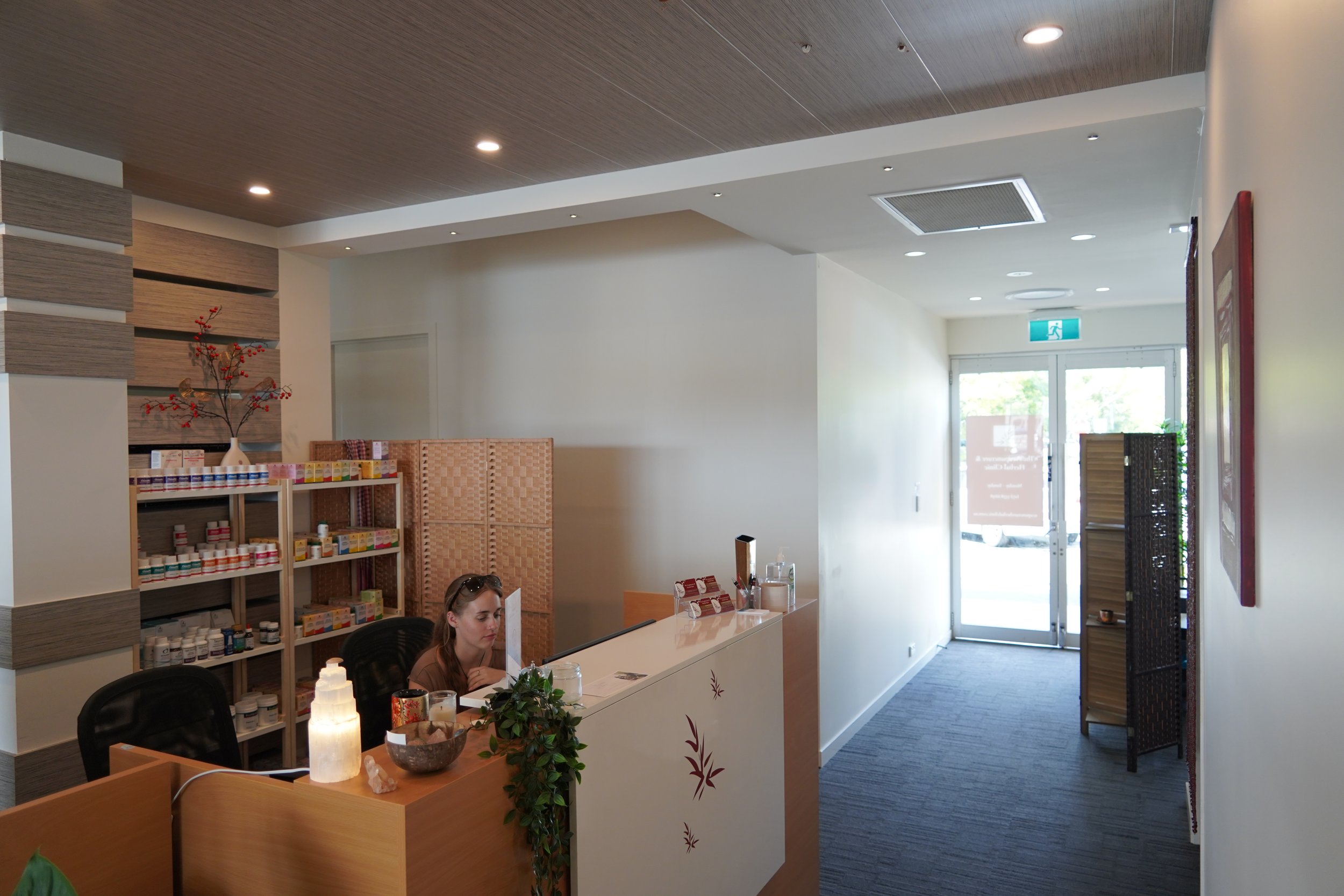 Reception area of a clinic or wellness center with a woman sitting at the counter, shelves with supplies, a decorative plant, and a privacy screen in the background.
