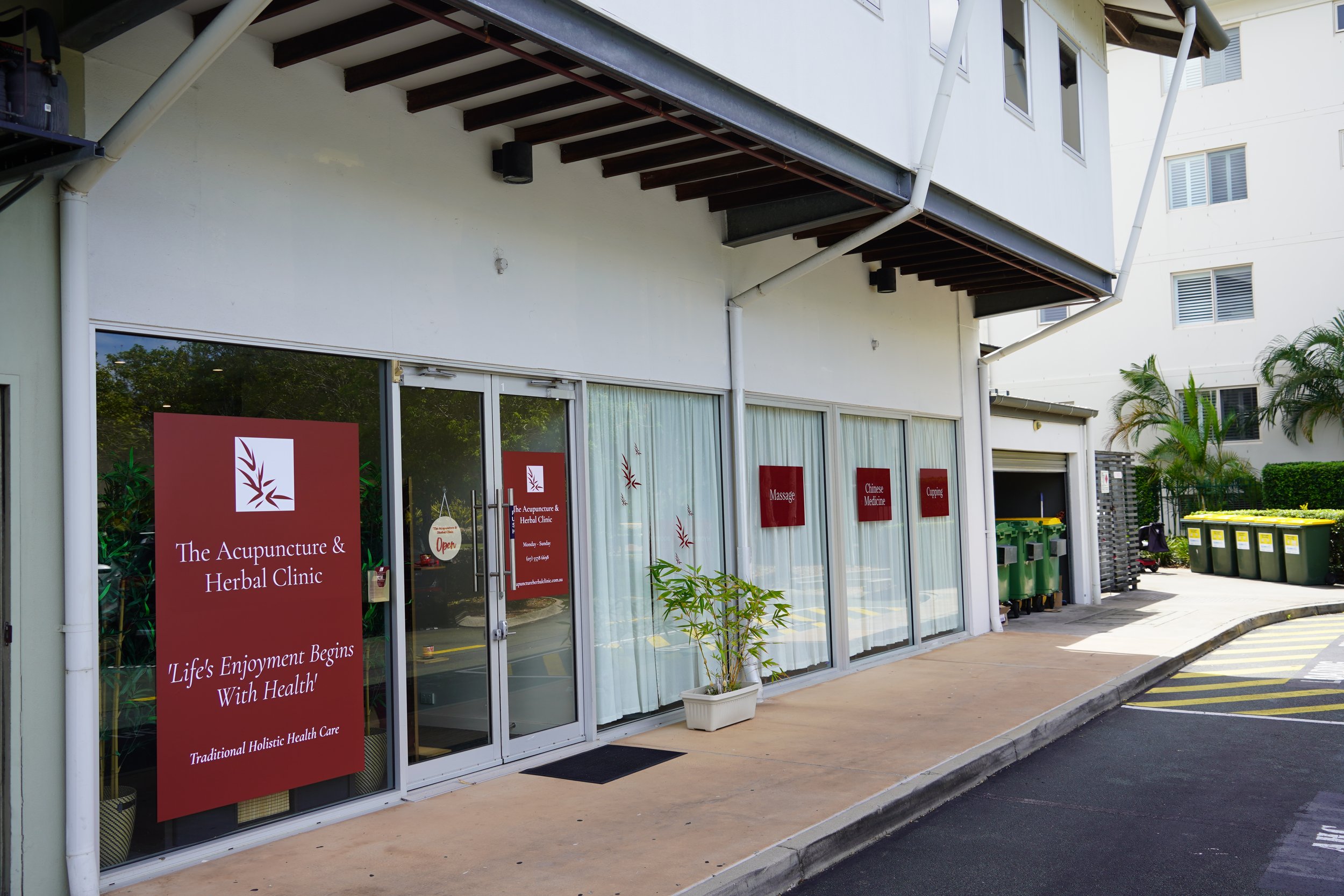 Exterior view of The Acupuncture & Herbal Clinic with large red signs on glass windows indicating services such as massage, Chinese medicine, and cupping. Green trash bins line the sidewalk.