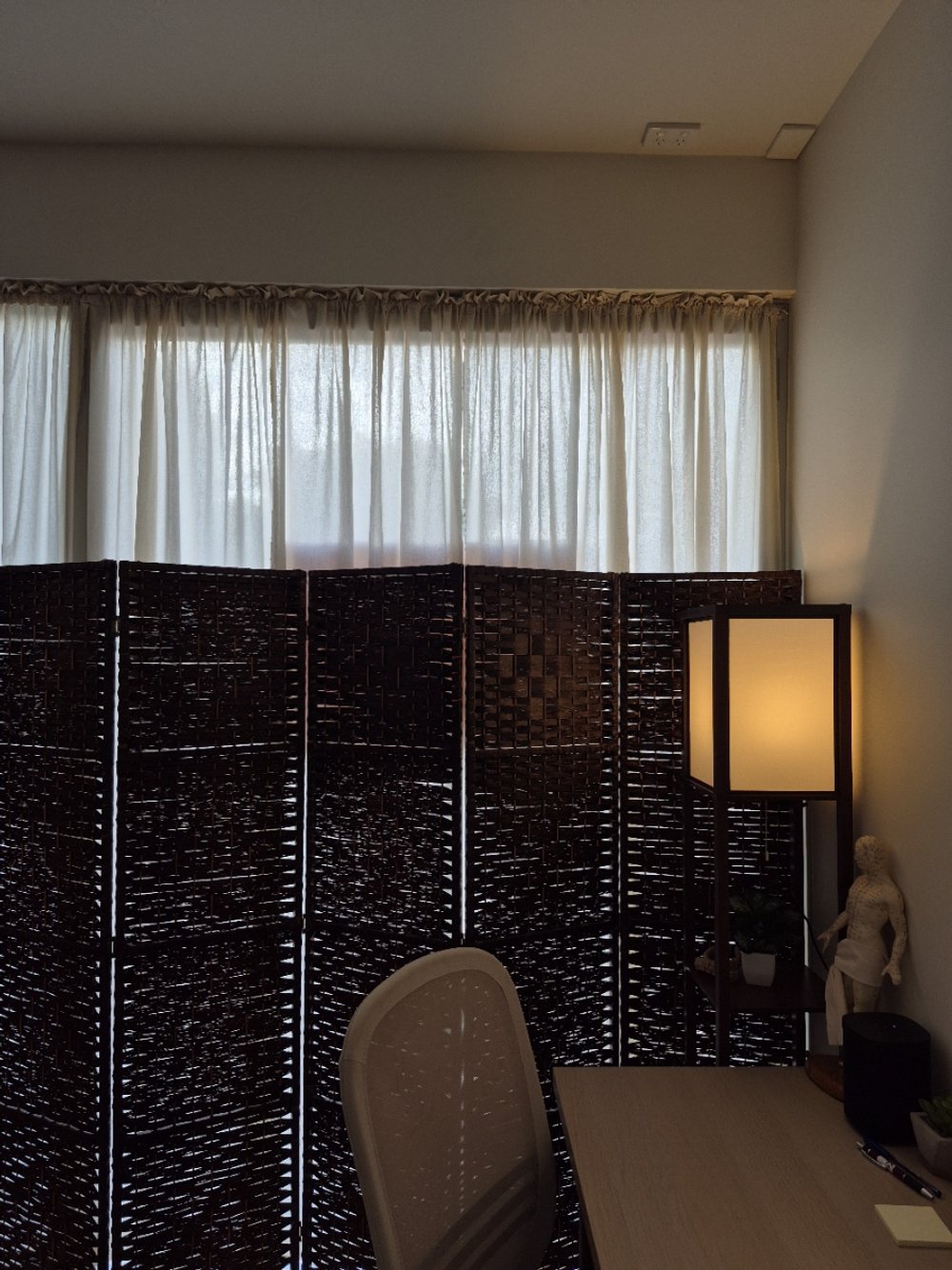 A room with white curtains, a dark woven folding screen, a beige chair, a lamp with a warm glow, and various decorative items on a shelf.