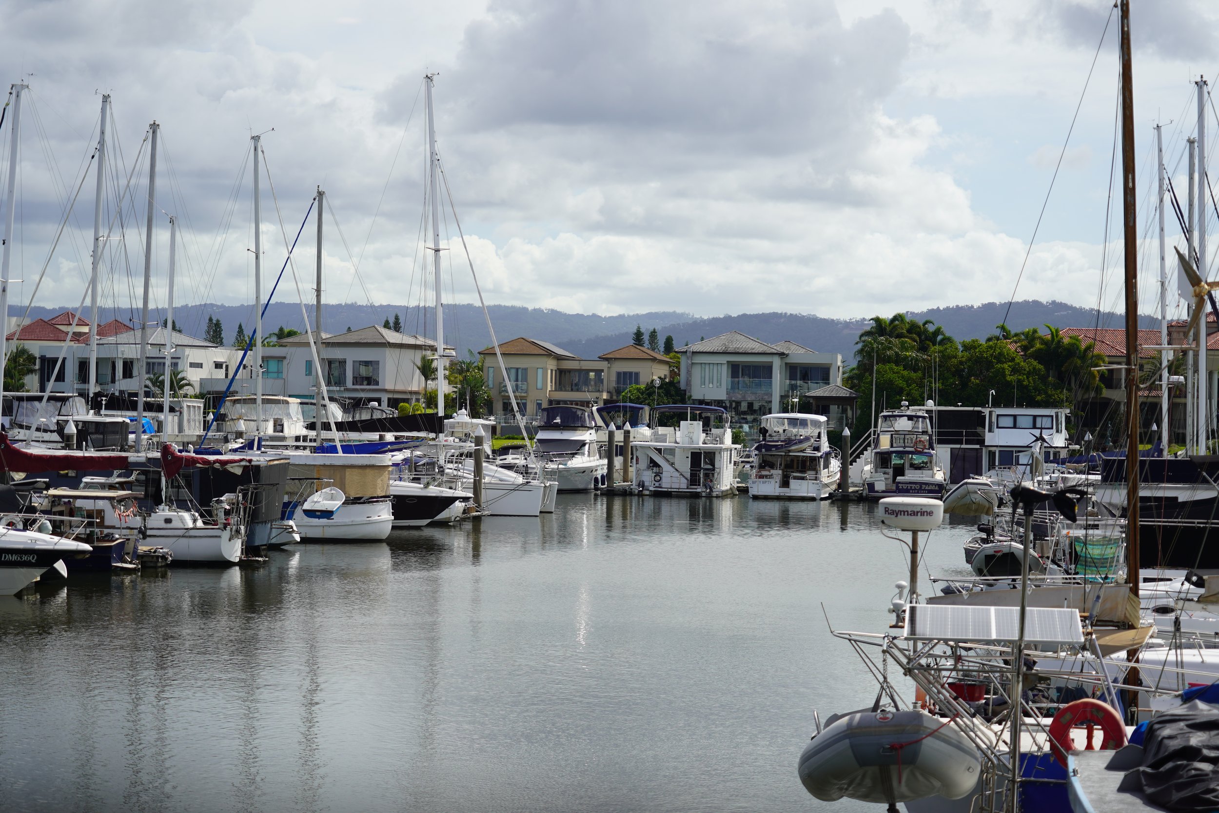 A marina with several boats docked, residential houses in the background, and mountains under a cloudy sky.