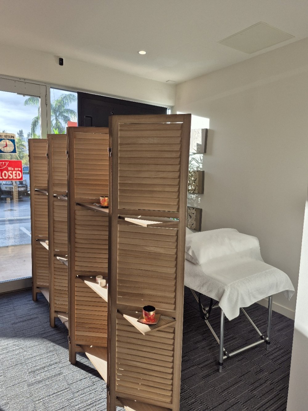 A room with a wooden privacy screen, a massage table covered with a white towel, and small wall shelves with decorative items, near front windows showing an outside view of palm trees and a parking lot.