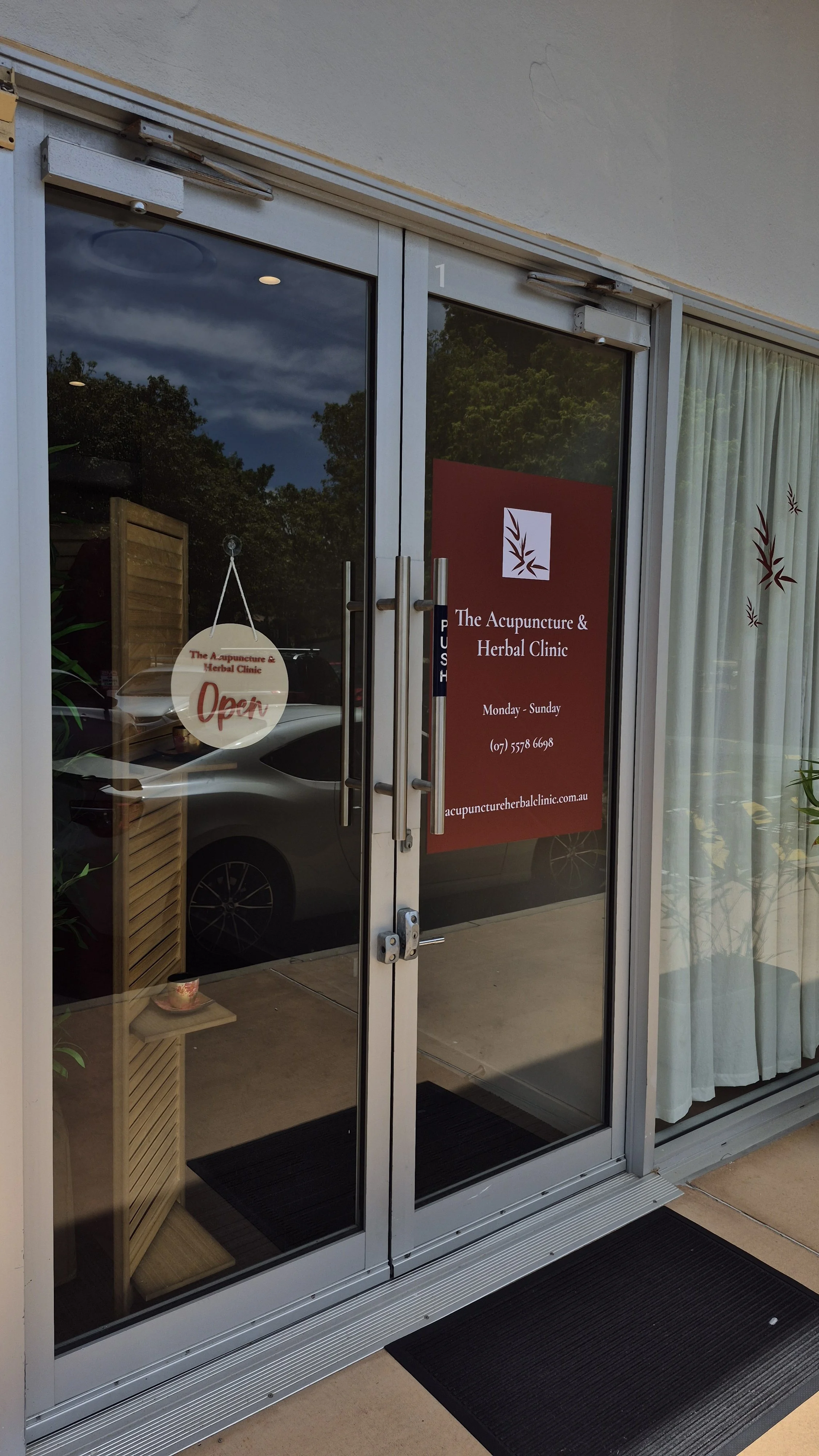 Glass door of The Acupuncture & Herbal Clinic with a red sign displaying hours, contact number, and website. An 'Open' sign hangs inside the door, and part of a parked car is visible through the glass.