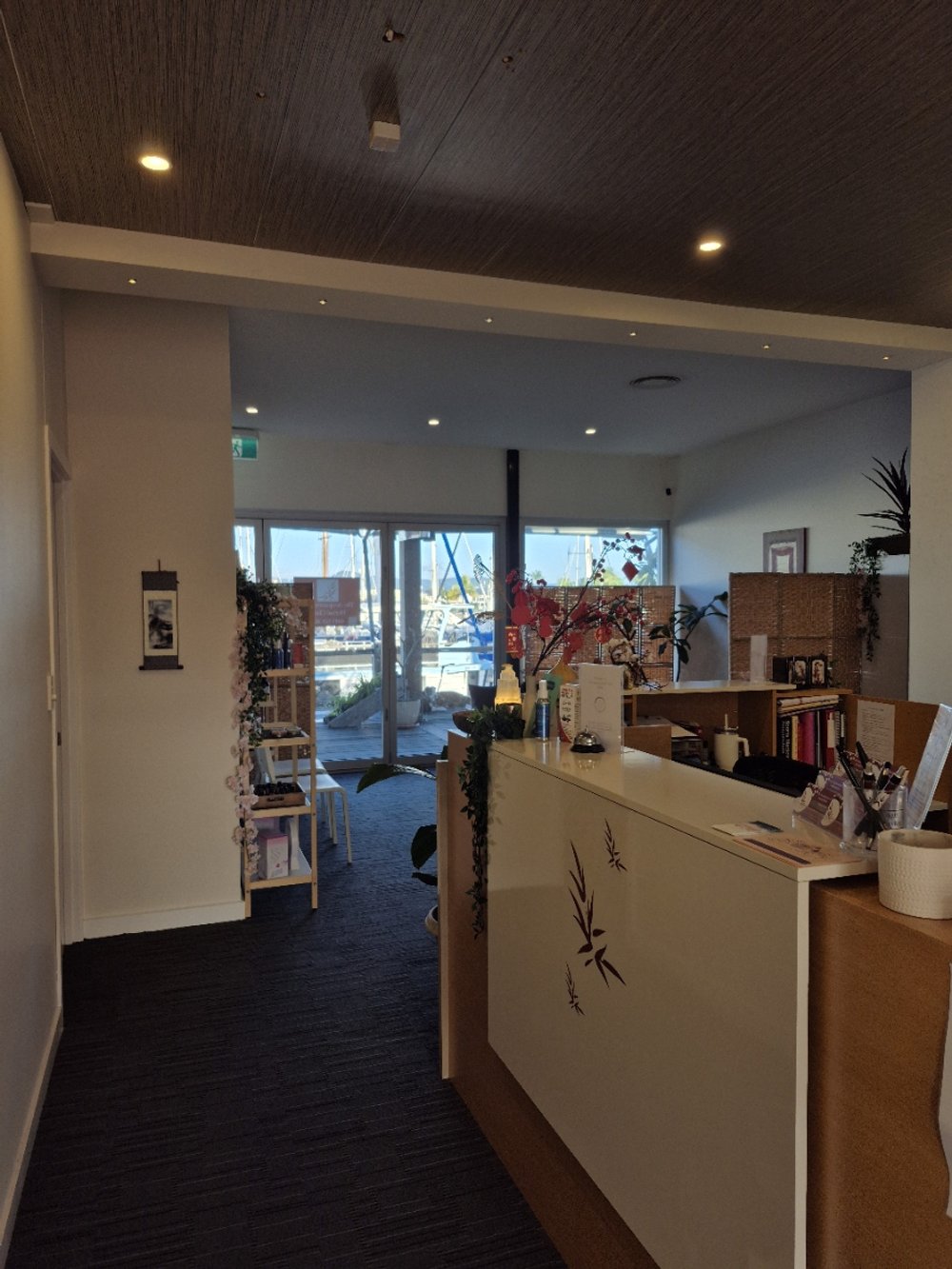 Interior of a cozy reception or small office area with a wooden ceiling, reception desk decorated with flowers, and large glass doors leading outside to a marina with boats.