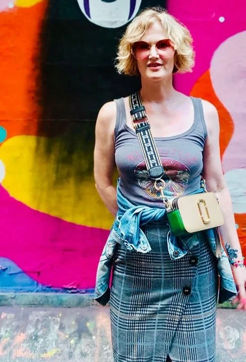 Woman (Helen Shearwood) with blonde curly hair wearing pink sunglasses, a gray sleeveless top, plaid skirt, and a crossbody bag, standing in front of a colorful graffiti wall.