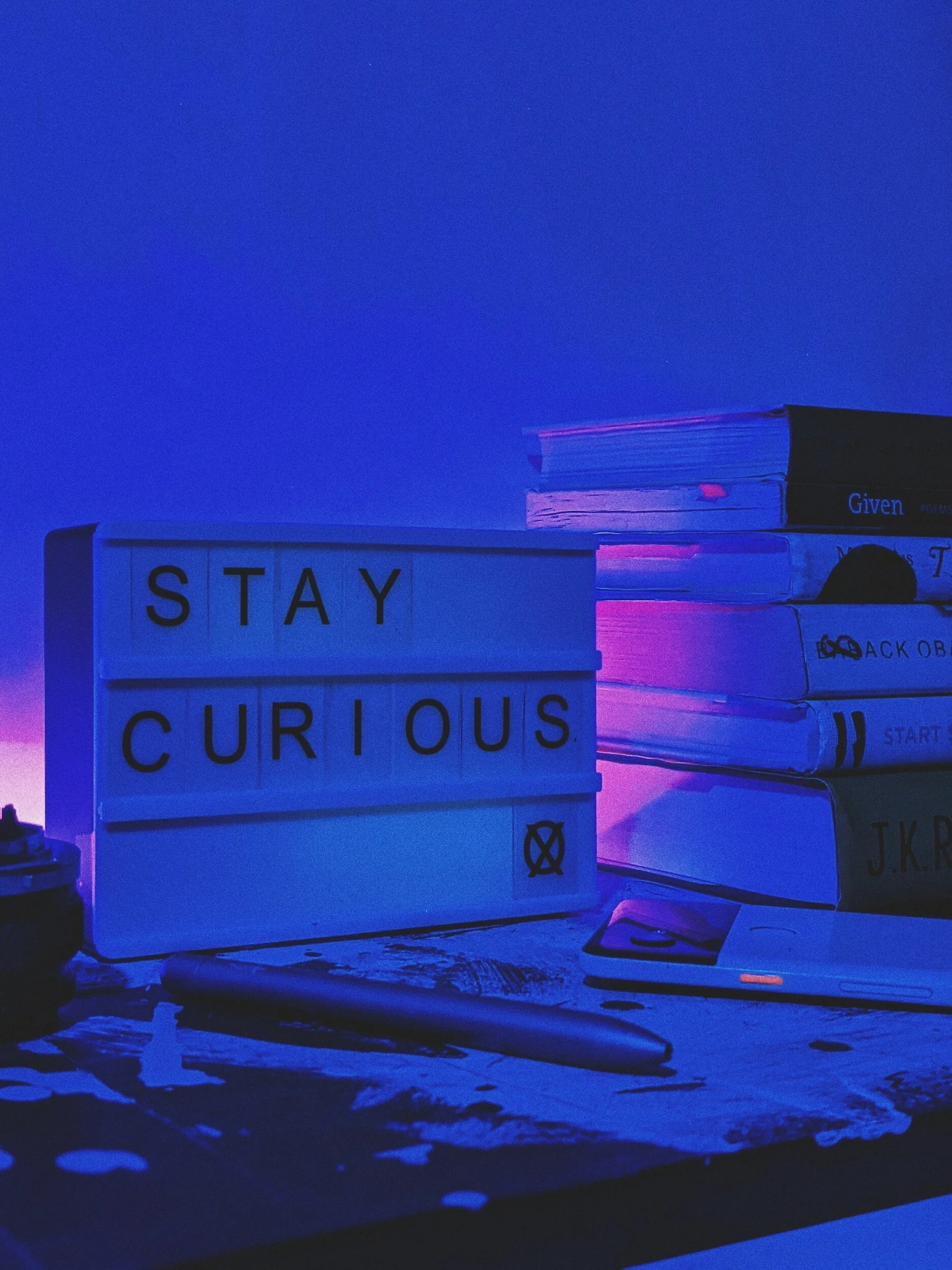 A lightbox sign with the words 'STAY CURIOUS' on a table with books, pen, paper, and a smartphone, all illuminated by blue and purple lighting.