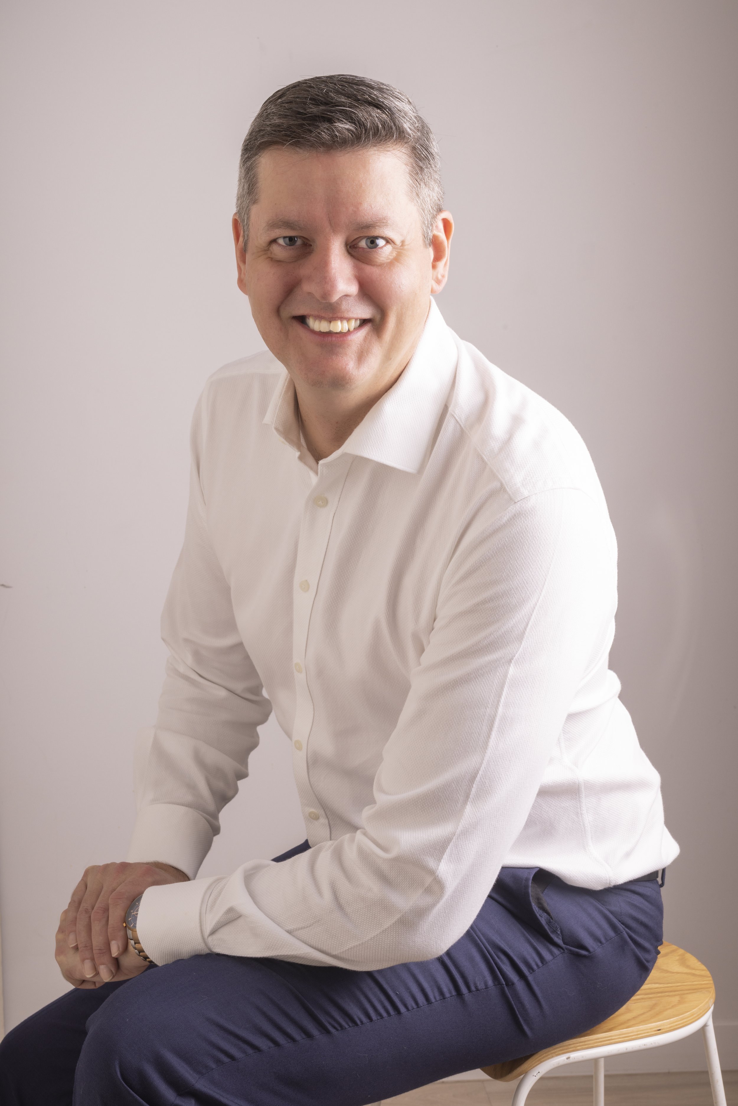 A smiling man in a white dress shirt and navy pants sitting on a wooden stool against a plain, light-colored background.