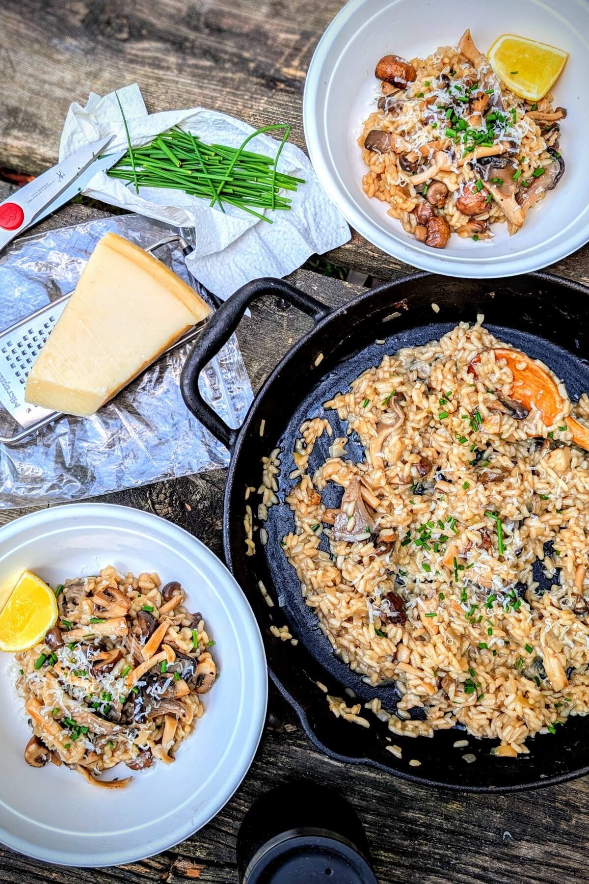 Two bowls and a cast iron pan of creamy mushroom risotto on a camp table with cheese and chives on the side