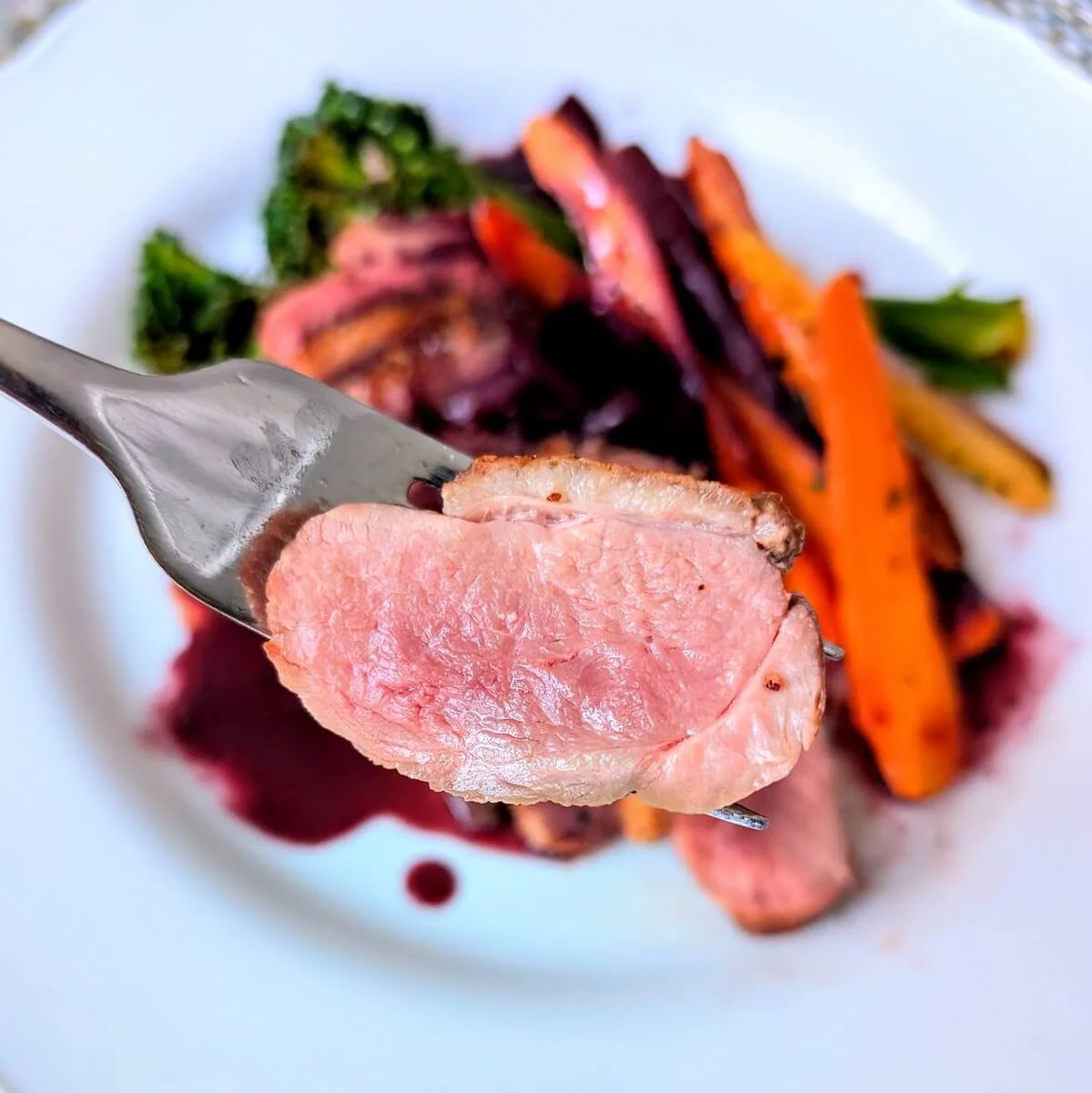 Perfectly cooked slice of crispy duck breast on a fork with duck pan sauce and vegetables in the background