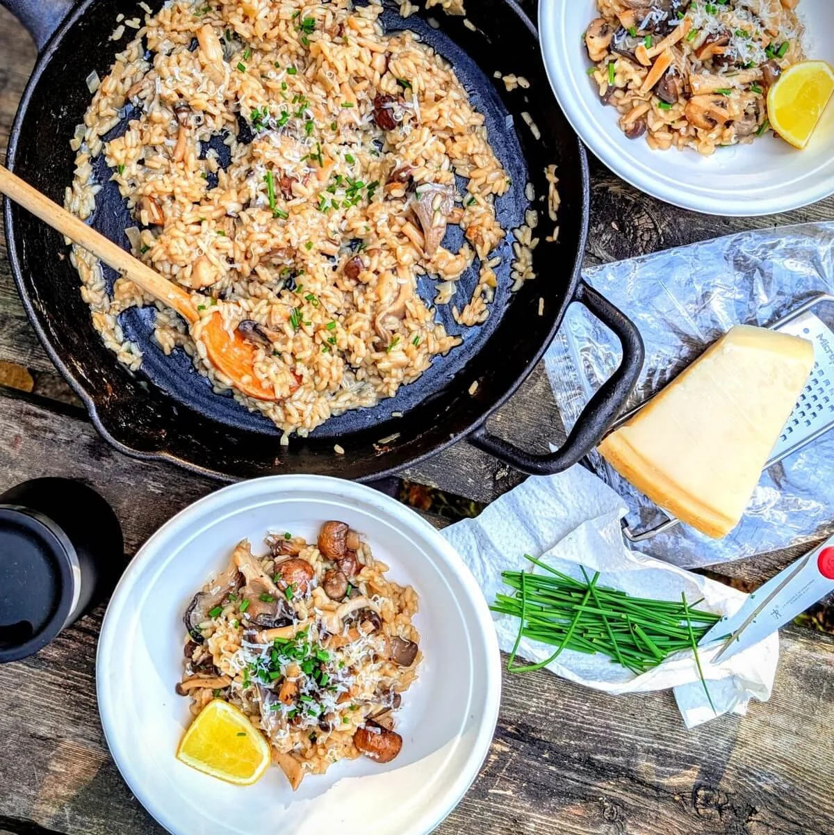 Camping table with two bowls of creamy mushroom risotto and a cast iron skillet with parmesan and fresh chives at the campsite
