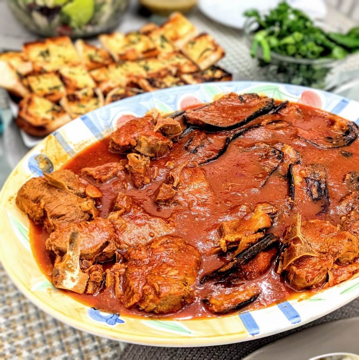 Platter of margat baytinjan, an Iraqi lamb and eggplant stew, served with garlic bread and fresh herbs in the background