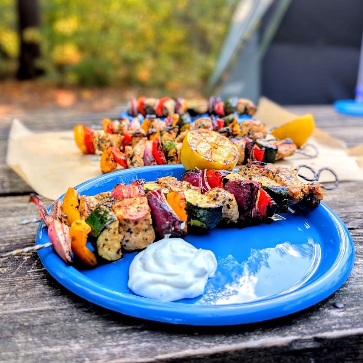Greek marinated chicken skewers served on blue plates at a campsite with tzatziki, charred lemon, and a tent in the background