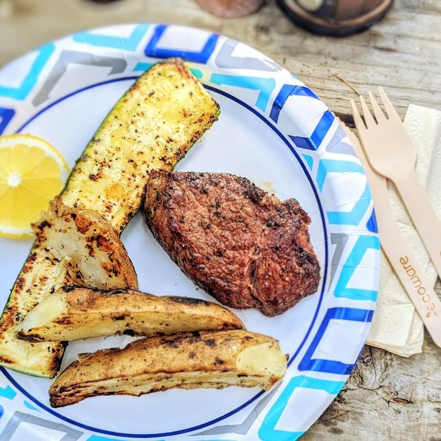 Disposable paper plate with grilled steak, potatoes, and bamboo cutlery on a camping picnic table