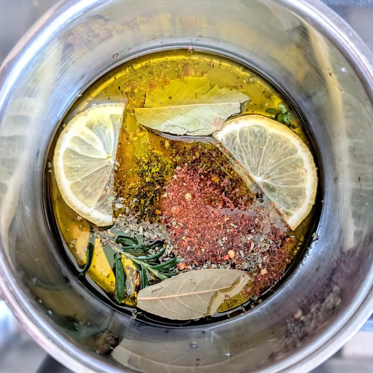 Lemon slices, chili flakes, rosemary, thyme, oregano, bay leaves, and olive oil in a pot for homemade marinated antipasto dressing
