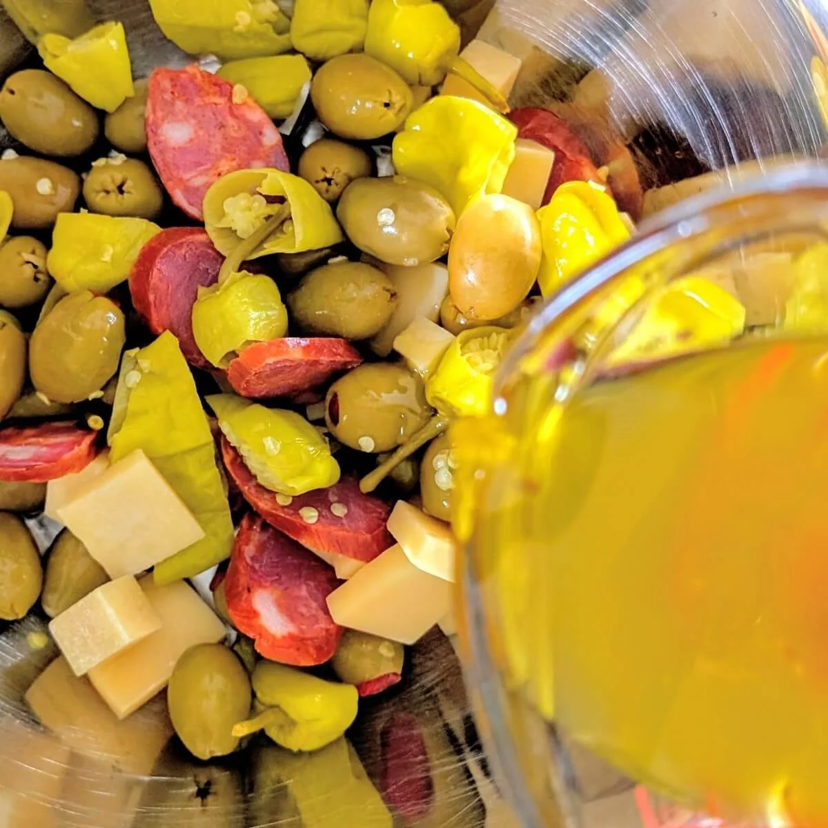 Antipasto marinade being poured into a bowl with olives cheese chorizo and pepperoncini peppers
