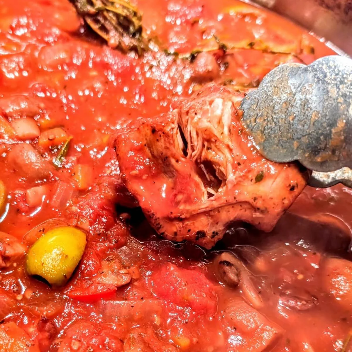 A pair of tongs lifting a piece of fall-apart turkey from a pot of simmered cacciatore