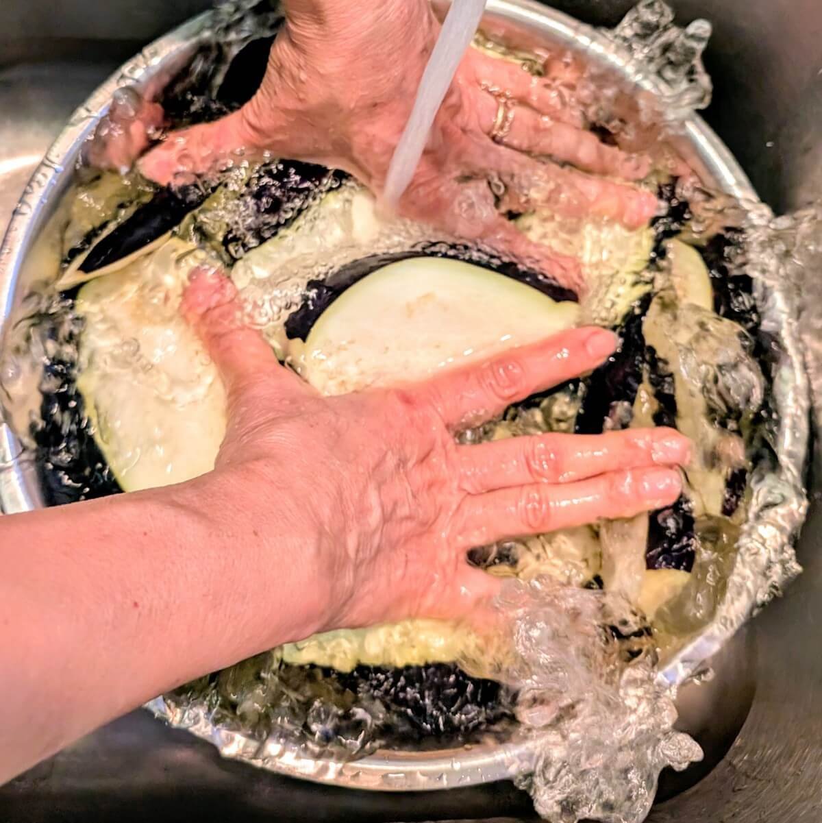 Hands pressing sliced eggplant down in a bowl of water while rinsing