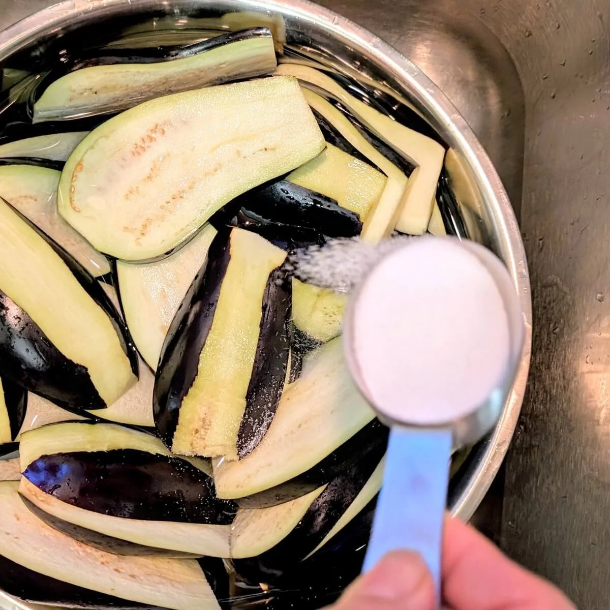 Salt being sprinkled into a bowl of sliced eggplant and water to create a simple brine