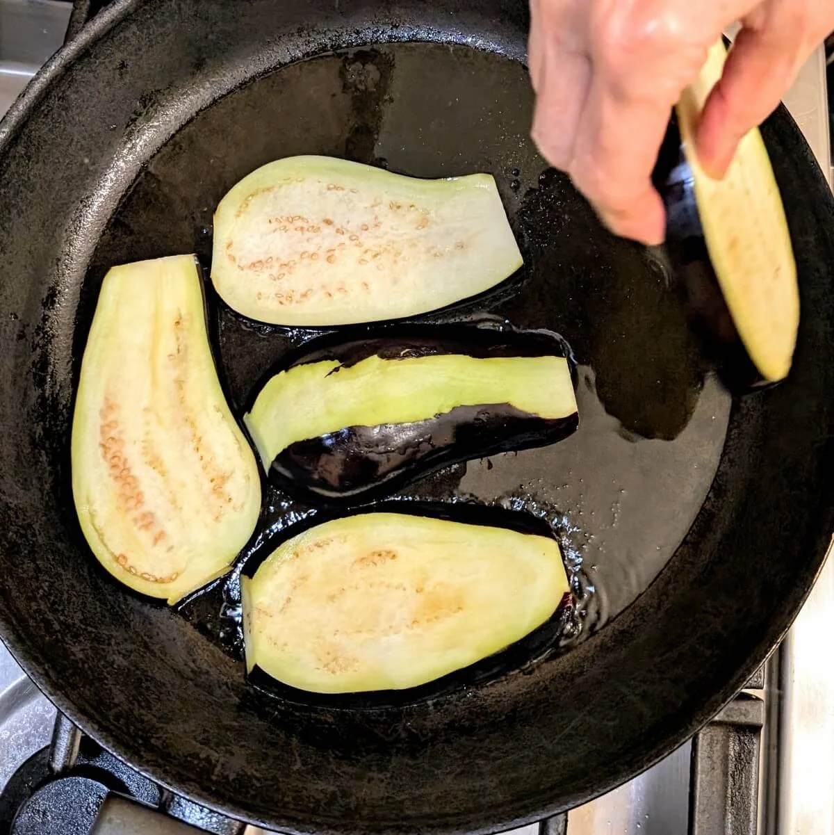 Sliced eggplant being placed into a hot skillet with olive oil