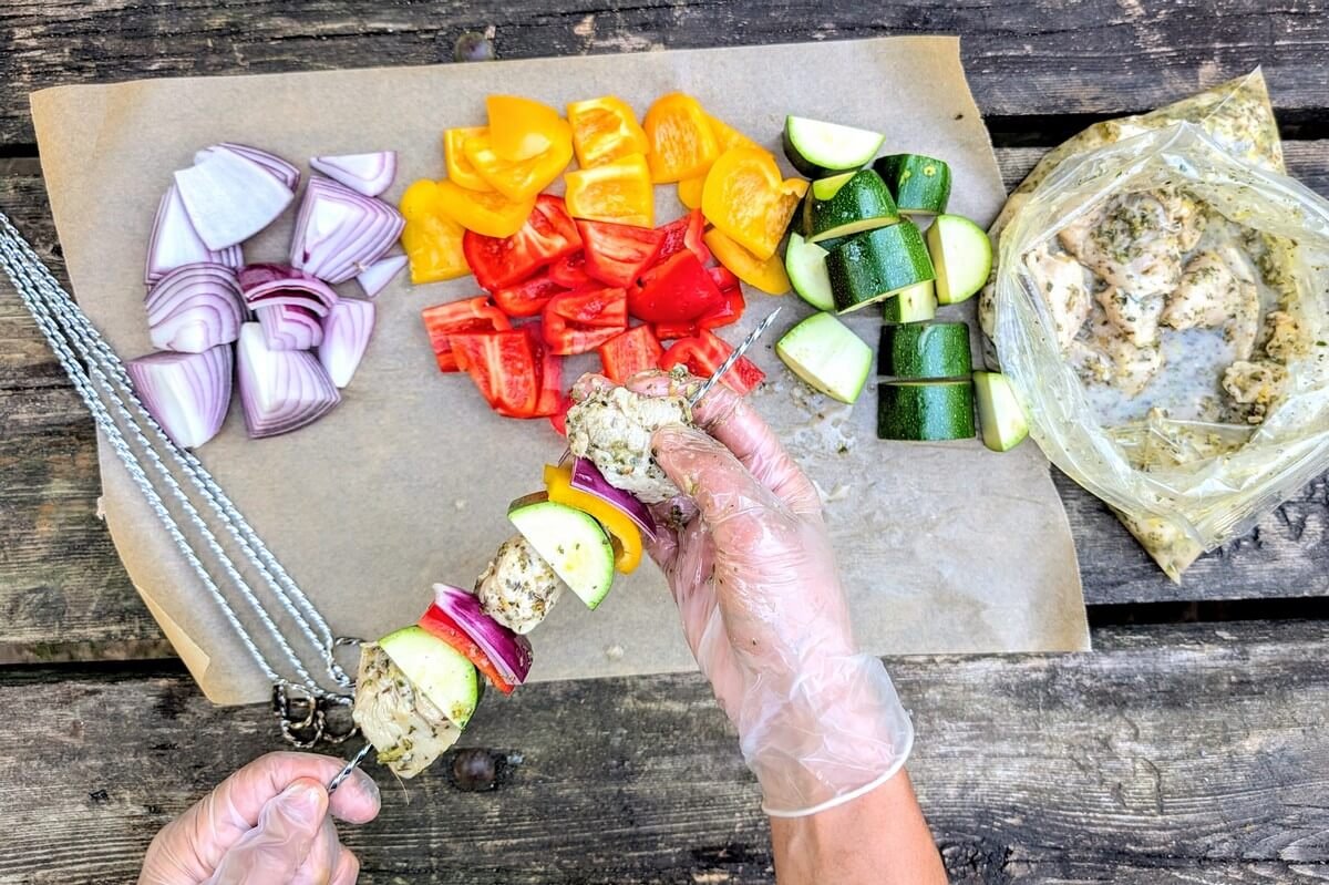 Gloved hands threading marinated chicken and vegetables onto skewers at a campsite