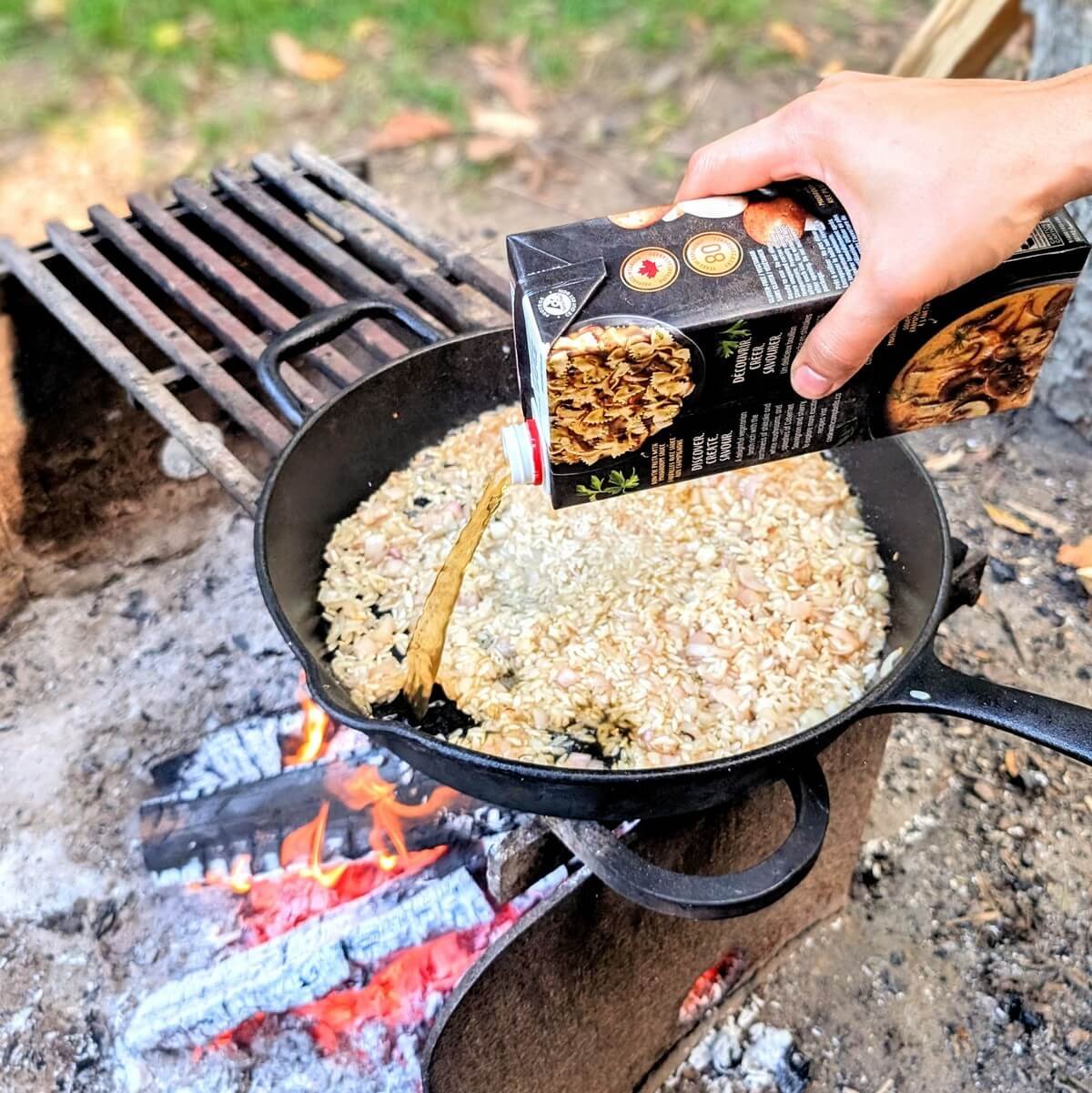 Hand pouring mushroom broth into arborio rice in a cast iron skillet over a campfire for campfire mushroom risotto