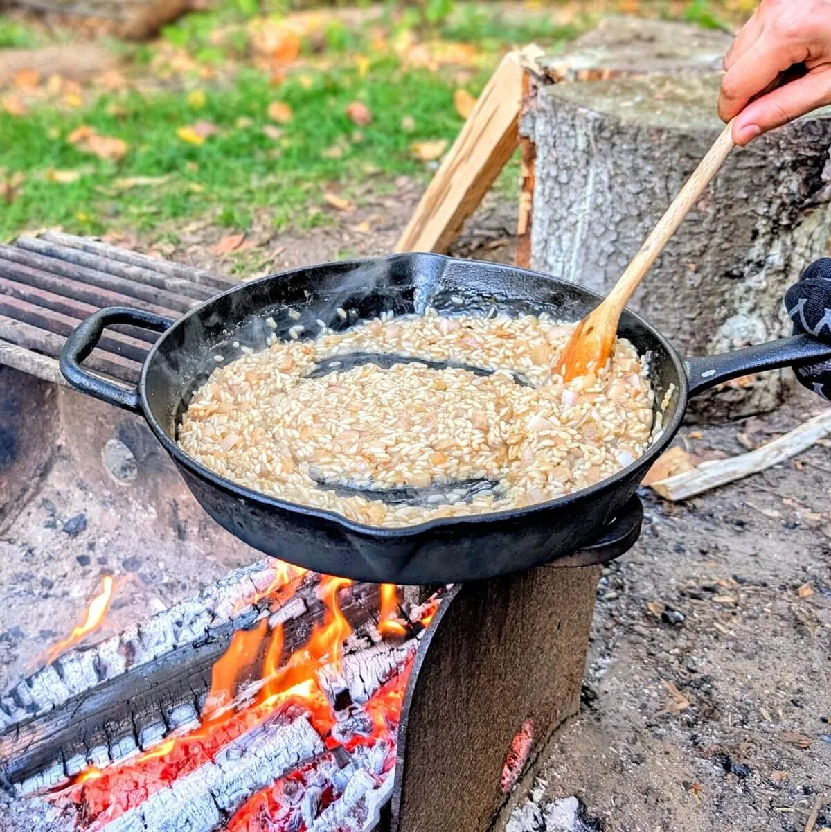 Stirring arborio rice with a wooden spoon as broth absorbs in cast iron pan over open fire for outdoor mushroom risotto