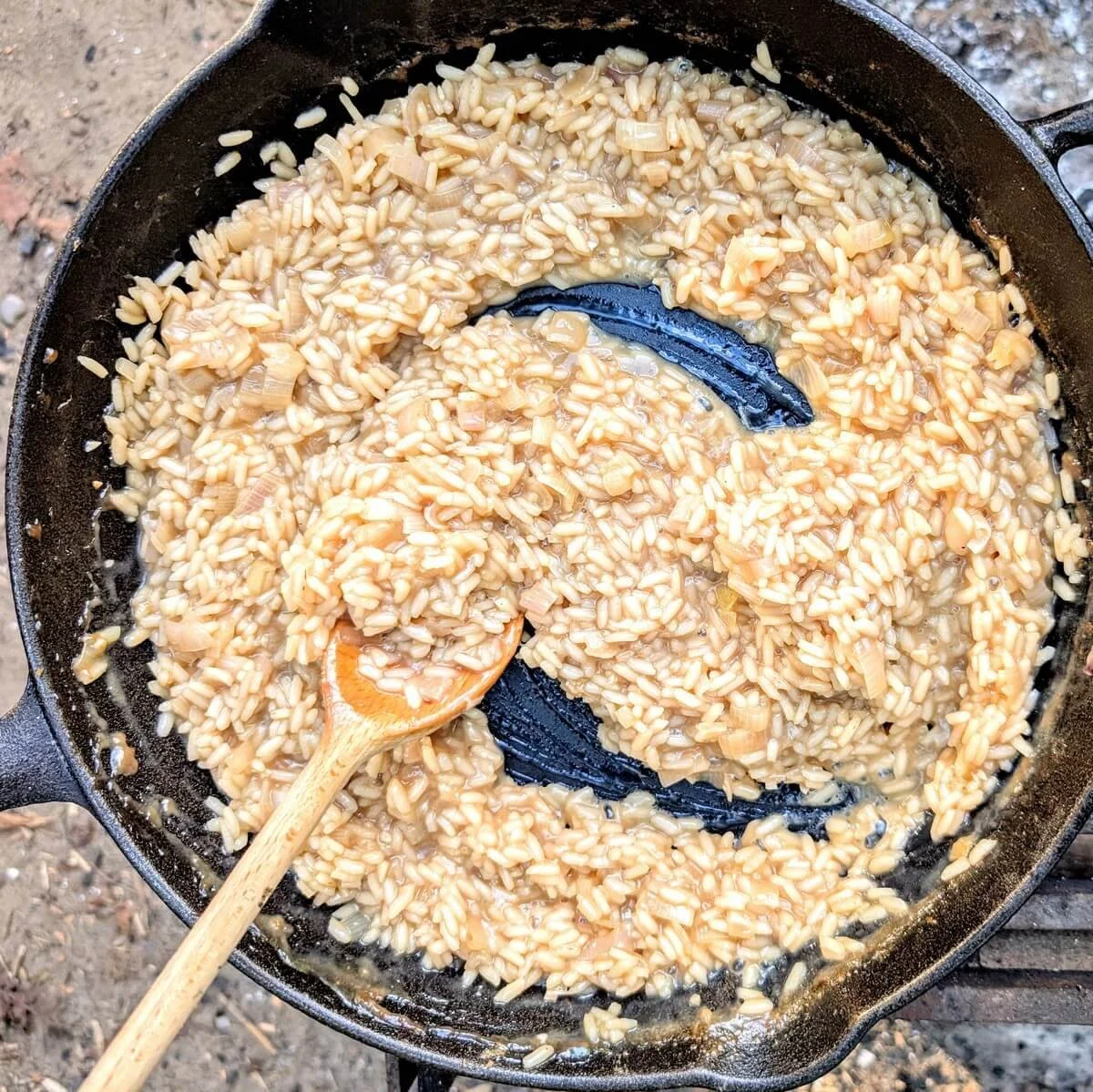 Creamy mushroom risotto cooking to al dente texture in cast iron pan over campfire flames