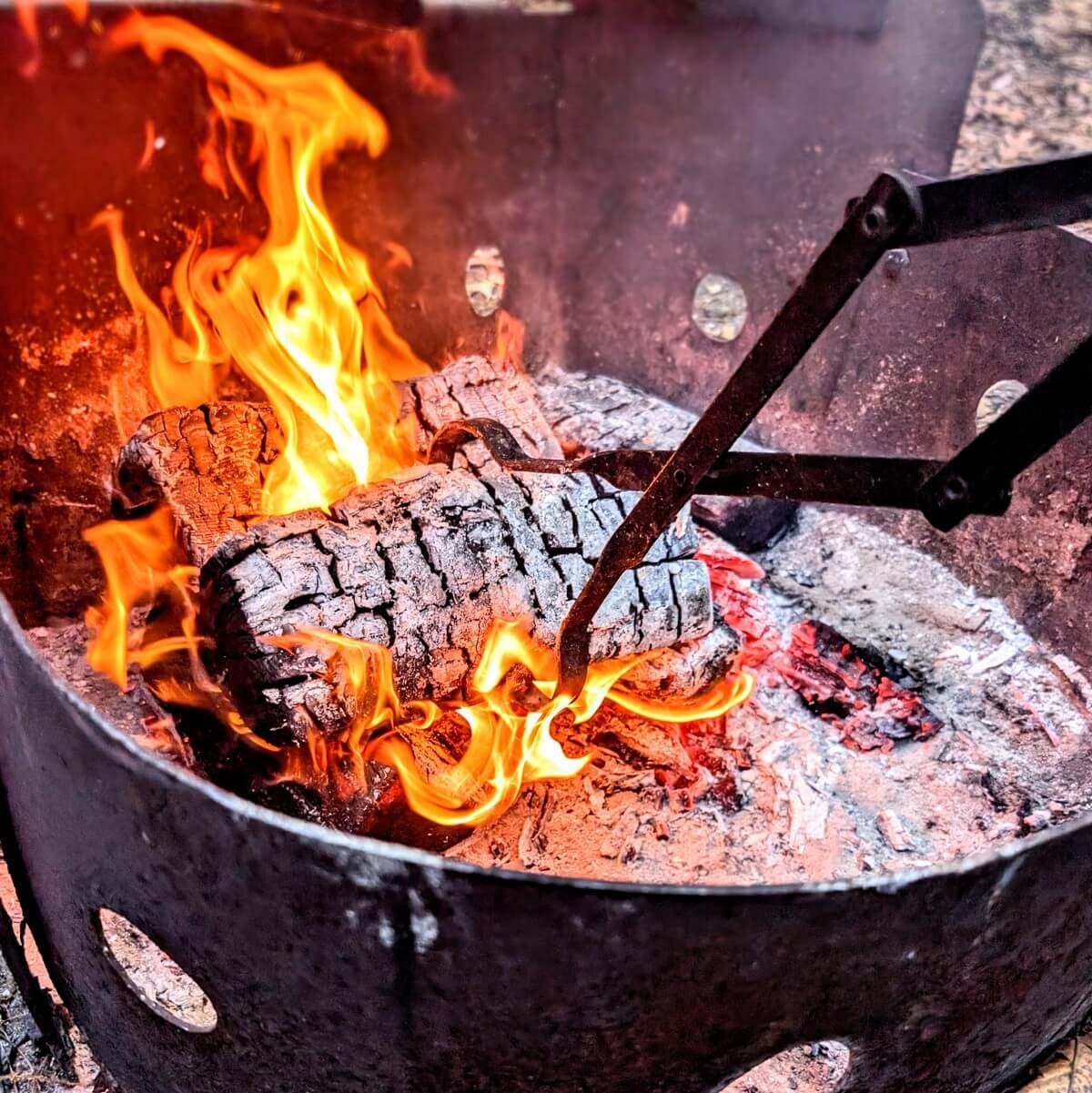 Firewood tongs gripping a burning log over bright flames inside a metal campfire ring