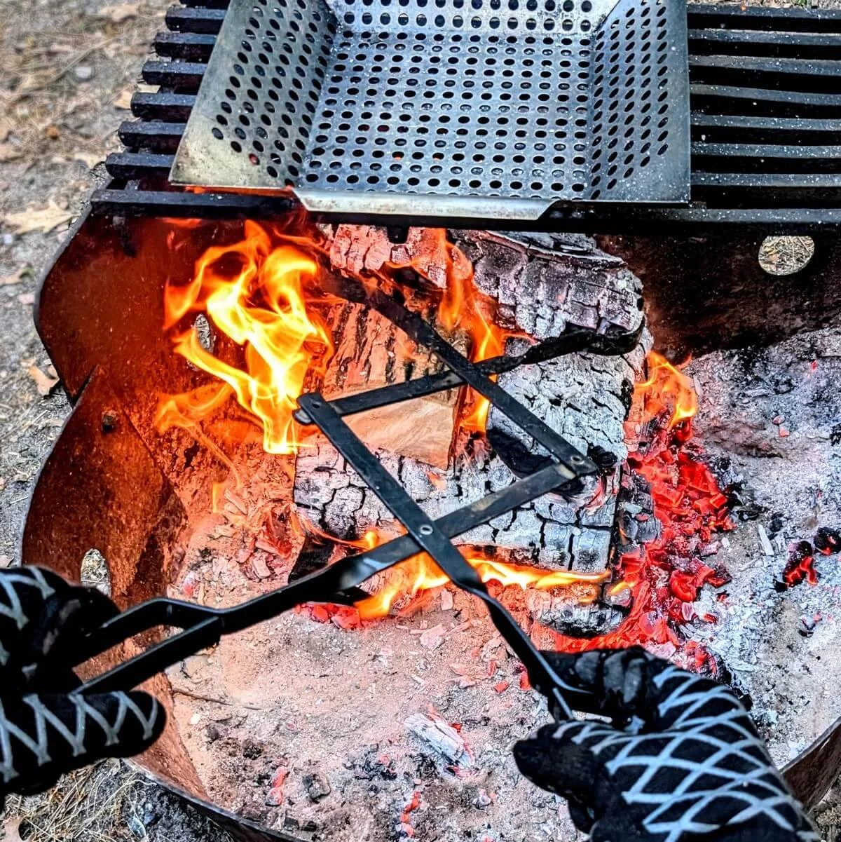 Two gloved hands using long steel firewood tongs to adjust burning logs and hot coals in a fire pit
