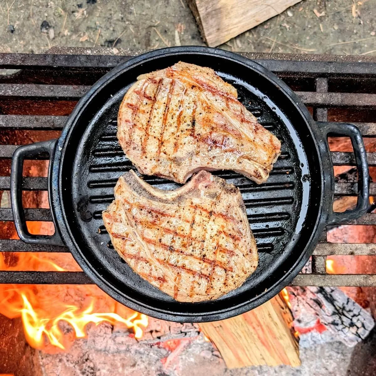 Cast-iron grill pan searing pork chops with grill marks over an open campfire